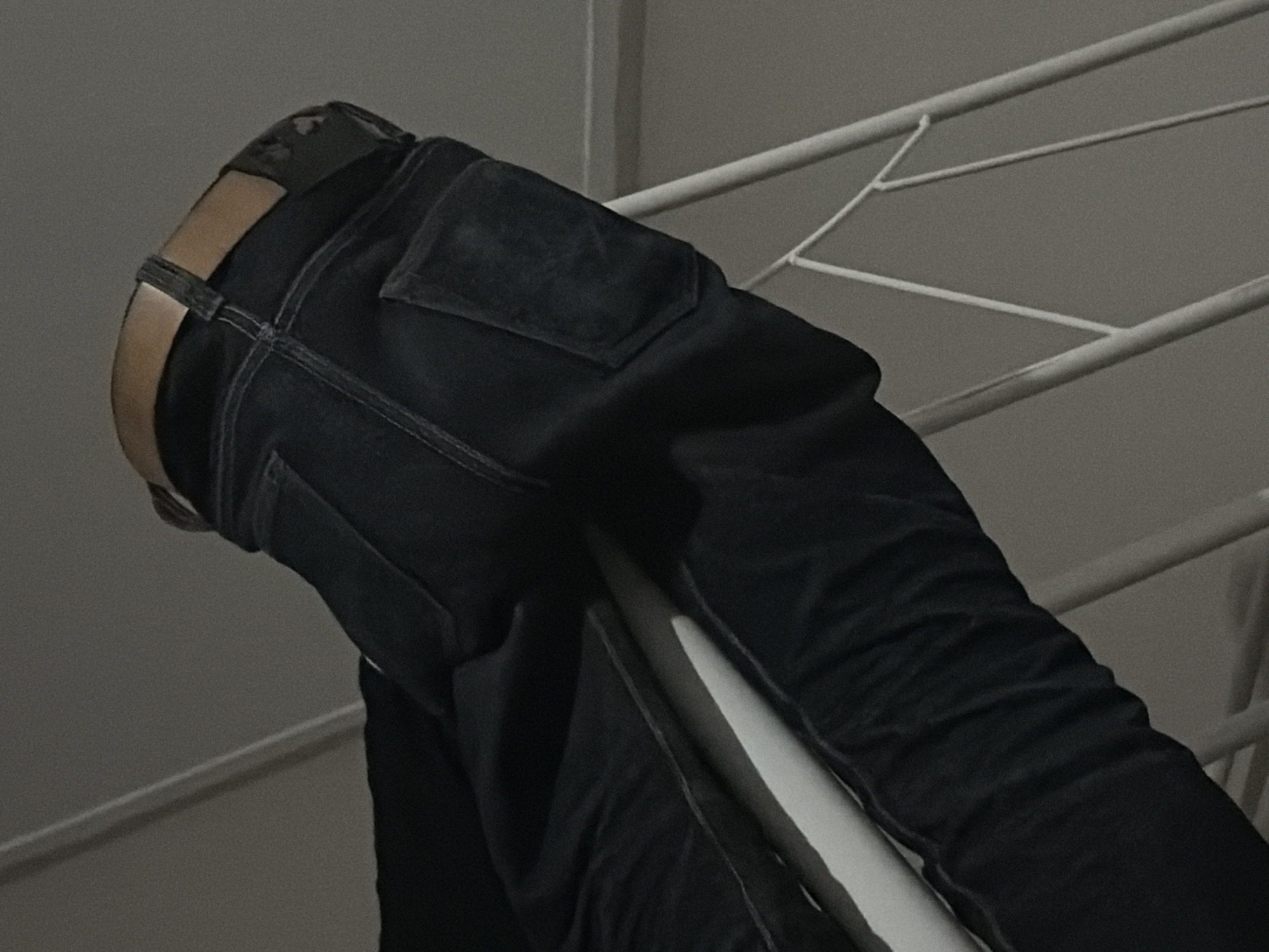 A low-angle, close-up shot depicts the rear view of a person wearing dark denim jeans and a brown leather belt. The person's legs and hips are visible, and they appear to be leaning or resting on a white metal railing. The jeans have a single back pocket visible on the right side, and the belt buckle is also partially obscured. The background consists of a plain, light-colored wall and the white metal structure. The scene is dimly lit, suggesting an indoor environment. No specific location in Tupah, Indonesia can be determined from the image.
