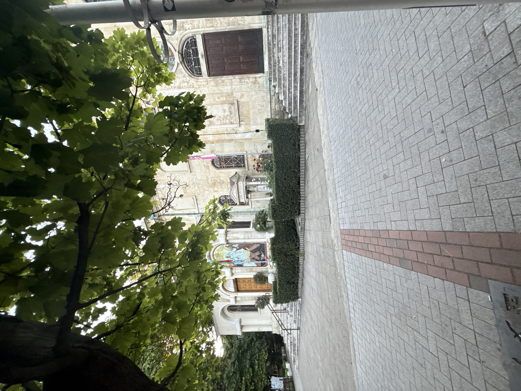 A street-level view of a church is captured under a canopy of green leaves from a tree in the foreground. The church is constructed of light-colored stone and features a prominent entrance with large wooden doors. A wide set of stone stairs leads up to the entrance. To the left of the main entrance, a smaller entrance is visible, also with wooden doors and stone stairs. A mosaic depicting religious figures adorns the facade to the right of the smaller entrance. Hedges are neatly trimmed in front of the church, and flowerbeds with red roses are present. The foreground consists of a paved courtyard with a distinct reddish-brown brick accent strip running through it. A metal pole stands on the right side of the image, partially obscuring the church facade. The sky is overcast.