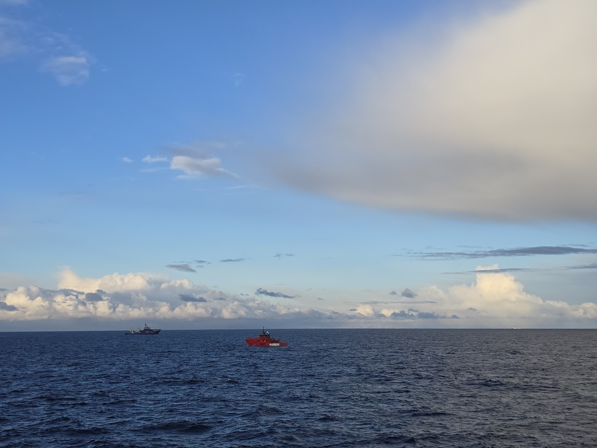 The image depicts a maritime scene under a partly cloudy sky. The sea is dark blue and moderately choppy. There are two ships visible on the horizon. One is a red ship, likely a service vessel. The other ship is a darker color and located further in the distance. The location is unknown.