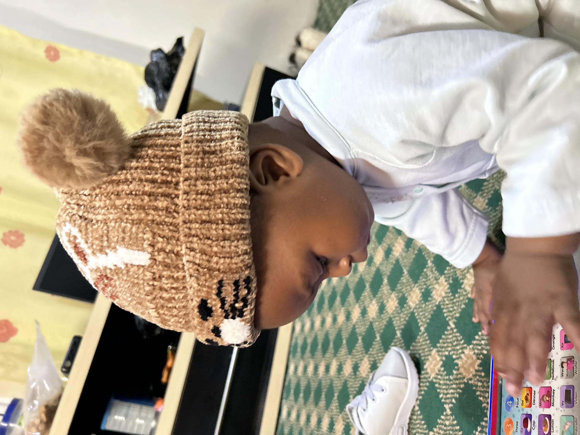 A young child, wearing a brown knitted hat with a pom-pom and a face design, is seated on a green and beige checkered carpet. The child is looking down at a children's electronic learning toy. A white sneaker is visible to the left of the child. In the background, a shelf unit holds various items, including bags and a black electronic device. The overall environment suggests an indoor setting, possibly a living room. The image is taken from a slightly elevated angle, focusing on the child and the toy.