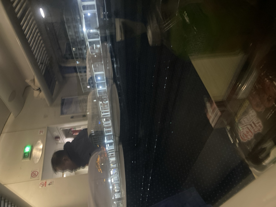 The image is a nighttime view inside a train car. A man in a dark jacket and white shirt is visible, likely using a restroom. The walls are white with various signs, including a green exit light and symbols. A window reflects the outside, showing a building at night. Dark, patterned seats are visible. Packaged food items rest on a table near the window, adding clutter to the reflection. The image has a blurred, layered effect due to the reflections.
