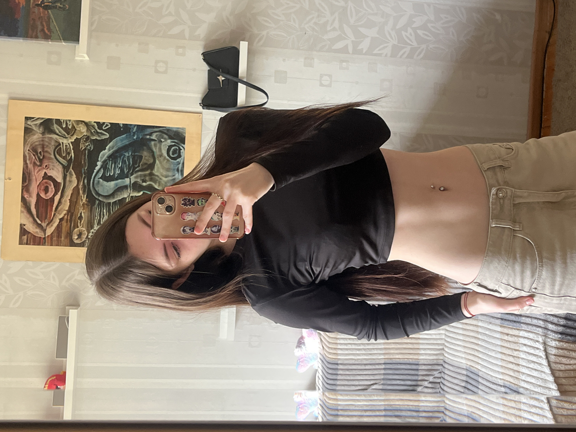 A young adult female is taking a mirror selfie in what appears to be a residential room in Unknown, Russia. She is wearing a black long-sleeved crop top and beige pants. The room has white wallpaper with a subtle leaf pattern. Behind her, there are two framed pictures on the wall, a black purse hanging on the wall, and a shelf with decorative items. The bottom half of the mirror reflects a striped bedspread. The woman is holding a phone with stickers on the back to take the photo. She has long brown hair and a double navel piercing.