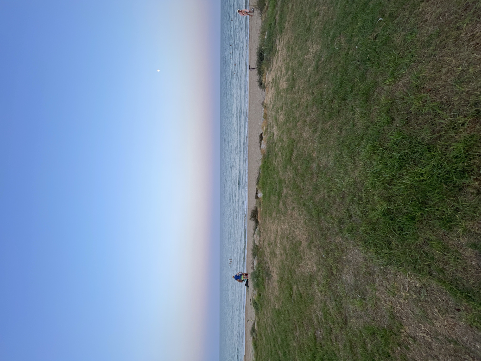 The image shows a serene beach scene at dusk, likely in Trikomo, Northern Cyprus. The sky transitions from a clear, deep blue at the top to soft hues of pink and purple near the horizon. A faint moon is visible in the upper right portion of the sky. The sea is calm with gentle waves rolling onto the sandy shore. In the foreground, a grassy area borders the beach. A few individuals are present on the beach and in the water. On the sand, a group of people appear to be sitting or standing, while others are swimming or wading in the shallow water. A woman in a straw hat and pink swimwear walks along the shoreline on the right side of the image. The overall atmosphere is peaceful and tranquil.
