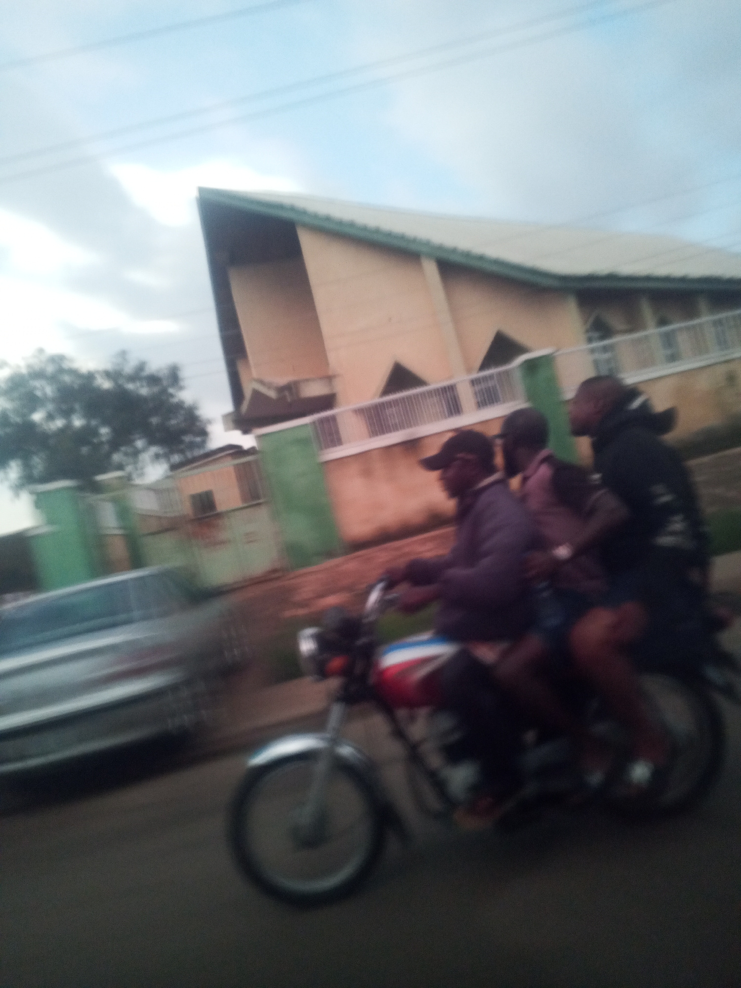 In an outdoor scene in Jos, Nigeria, three individuals are riding on a motorcycle. The motorcycle is moving on a road, indicated by the motion blur of the vehicle and the ground. The rider in the front is wearing a dark baseball cap and a purple jacket. The person in the middle is wearing a dark jacket over a lighter-colored shirt, and the person in the back is wearing a dark hooded jacket. They are traveling past a building with a light-colored facade and a green roof. The building has an angular architectural design. In front of the building is a light-colored railing and some green walls. To the left of the motorcycle, a silver car is visible, also exhibiting motion blur. The sky above is partly cloudy. The overall impression is of a busy street scene in the city.