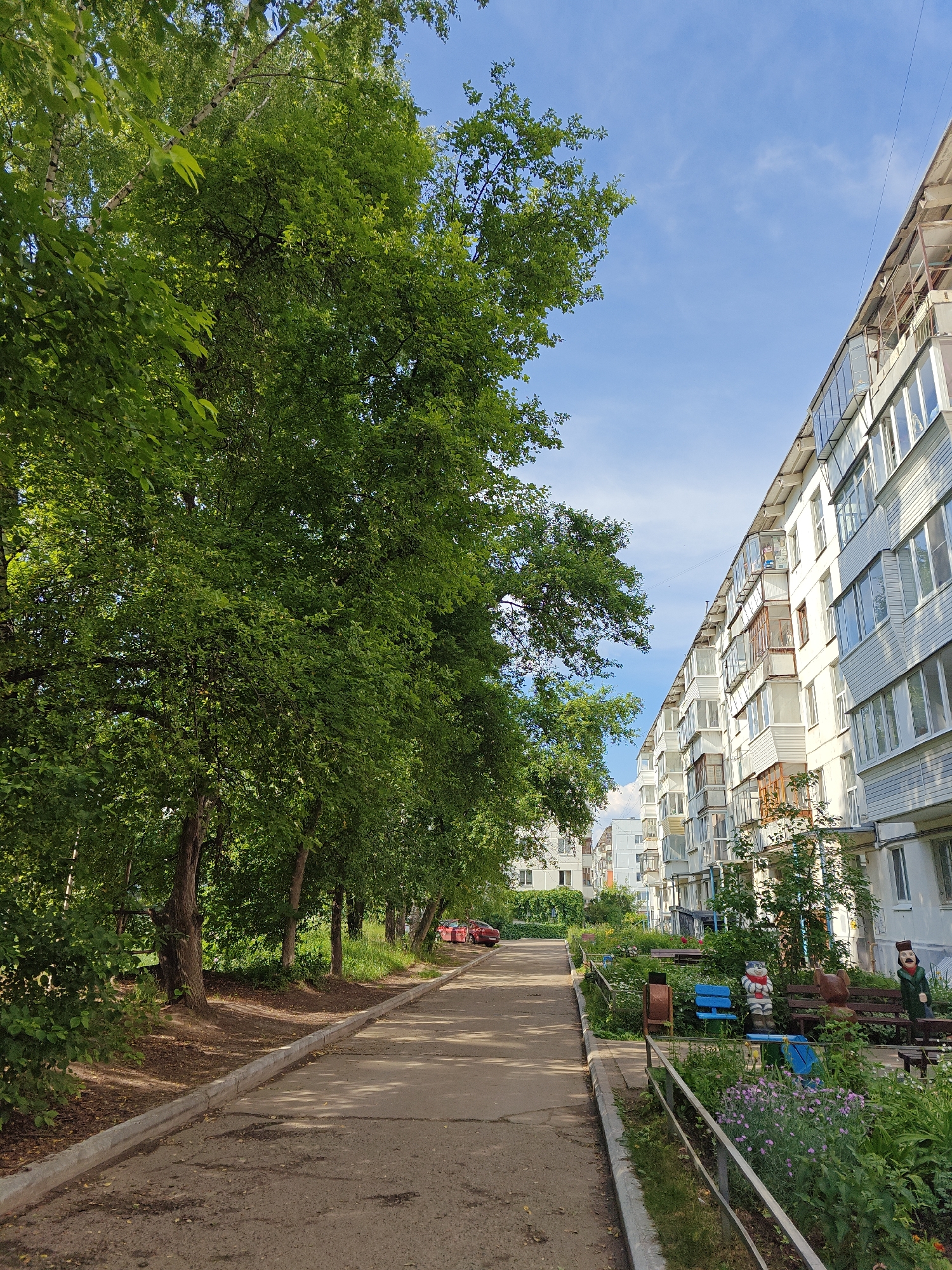 This is an outdoor, eye-level shot of a residential area in Chaikovsky, Russia. On the right, a multi-story apartment building stretches along a paved walkway lined with a metal fence. The building has numerous balconies and windows, with some residential activity visible through the glass. A small, fenced-in garden area with benches and decorative figures is situated between the walkway and the building. On the left, a row of mature trees with lush green foliage casts dappled shadows on the walkway. The walkway extends into the distance, where more buildings and trees are visible. The sky is clear blue with a few wispy clouds. A red car is parked further down the walkway on the left. The scene appears to be a quiet, everyday street in a Russian city, likely a residential courtyard during daylight hours.