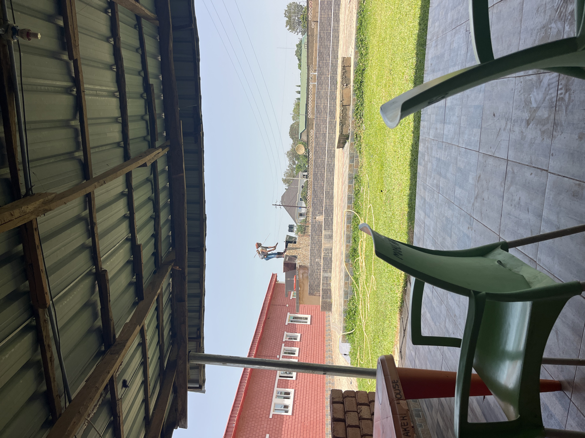 This media file captures an outdoor scene, likely in Jos, Nigeria, during daylight hours with clear skies, suggesting fair weather. The viewpoint is from under a rustic, unfinished tin roof structure, looking out onto a yard.
In the foreground, partially visible are the legs of green plastic chairs and the edge of an orange table. The table has faint text that appears to say "BAKER HOUSE".
Beyond the covered area, a brick building with a red roof and white-framed windows stands prominently. In the mid-ground, a person is seen working on a haystack. This individual, dressed in blue jeans and a light-colored shirt, appears to be using a tool, possibly a rake or pitchfork, to adjust the hay. The activity suggests agricultural work or preparation.
The background shows a neatly manicured green lawn, a brick pathway, and more buildings in the distance, along with trees and power lines, indicative of a semi-urban or rural setting. The overall impression is one of a functional, perhaps agricultural or hospitality establishment, with ongoing activities. No specific emotions are discernible from the image.