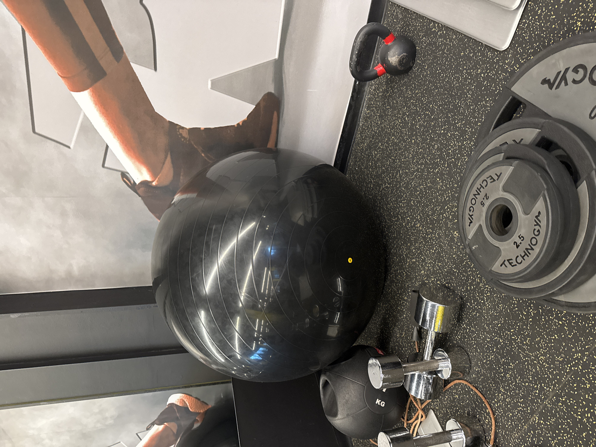 The image shows a collection of gym equipment placed on a dark, speckled rubber floor. Equipment includes a black exercise ball, dumbbells, kettlebell, and weight plates labeled "TECHNOGYM" and "2.5 KG." Part of a poster featuring a person's leg is also visible. The scene appears to be inside a gym, likely in Sofia, Bulgaria, given the context and the brand of equipment, "TECHNOGYM", is commonly found in gyms across the city. No people are directly visible in the frame.