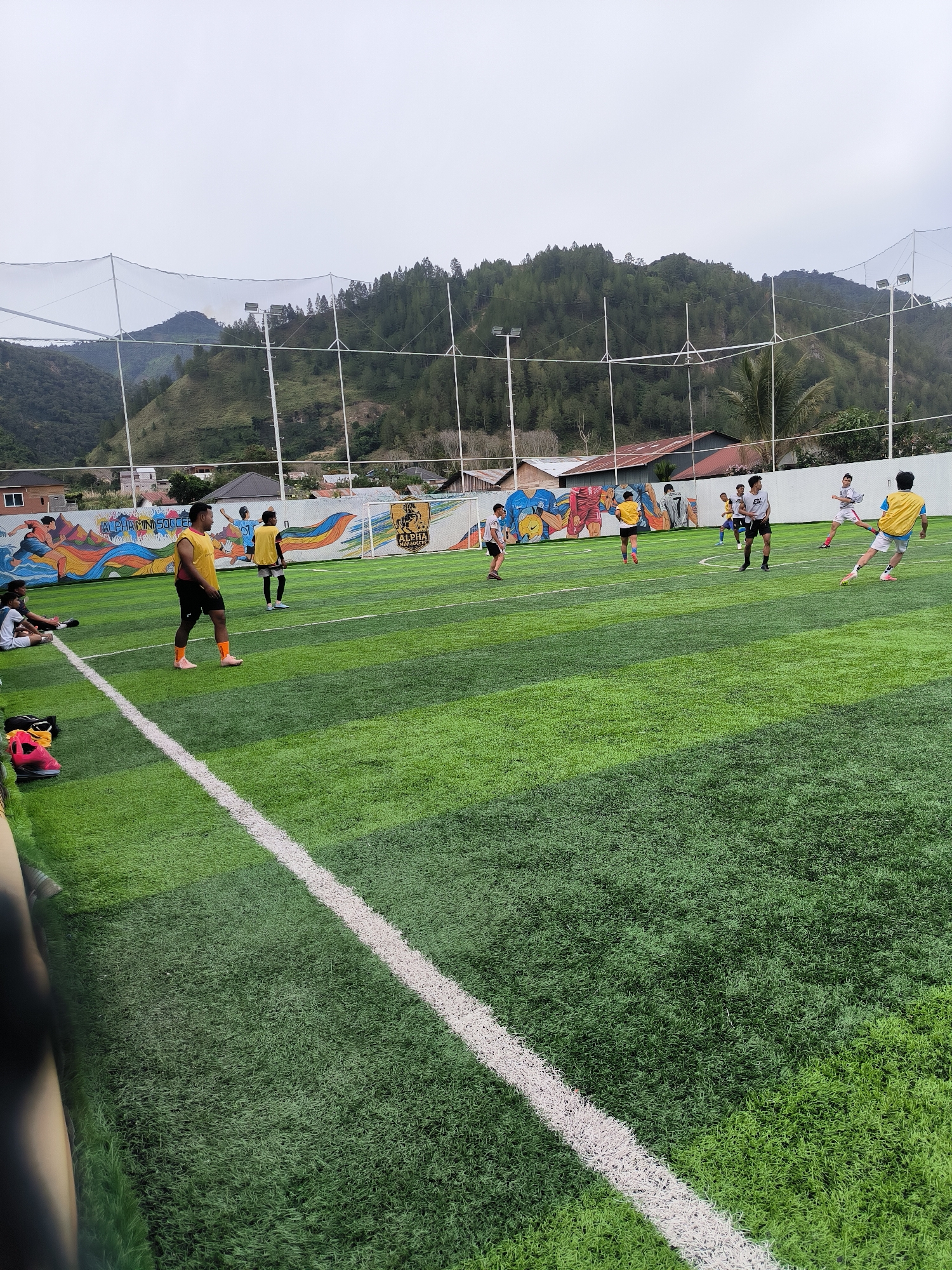 The image depicts an outdoor artificial turf soccer field in Mendale, Indonesia. Several individuals, appearing to be young men and boys, are engaged in a soccer game. Some are wearing yellow jerseys, while others are in white or grey. The field is marked with white lines and has a net enclosure. In the background, a mural with colorful imagery and the text "ALPHA MINI SOCCER" is visible on a wall. Beyond the field, there are buildings with corrugated metal roofs and lush green hills covered in trees under a cloudy sky. The overall scene captures a recreational soccer activity in a scenic, hilly location.