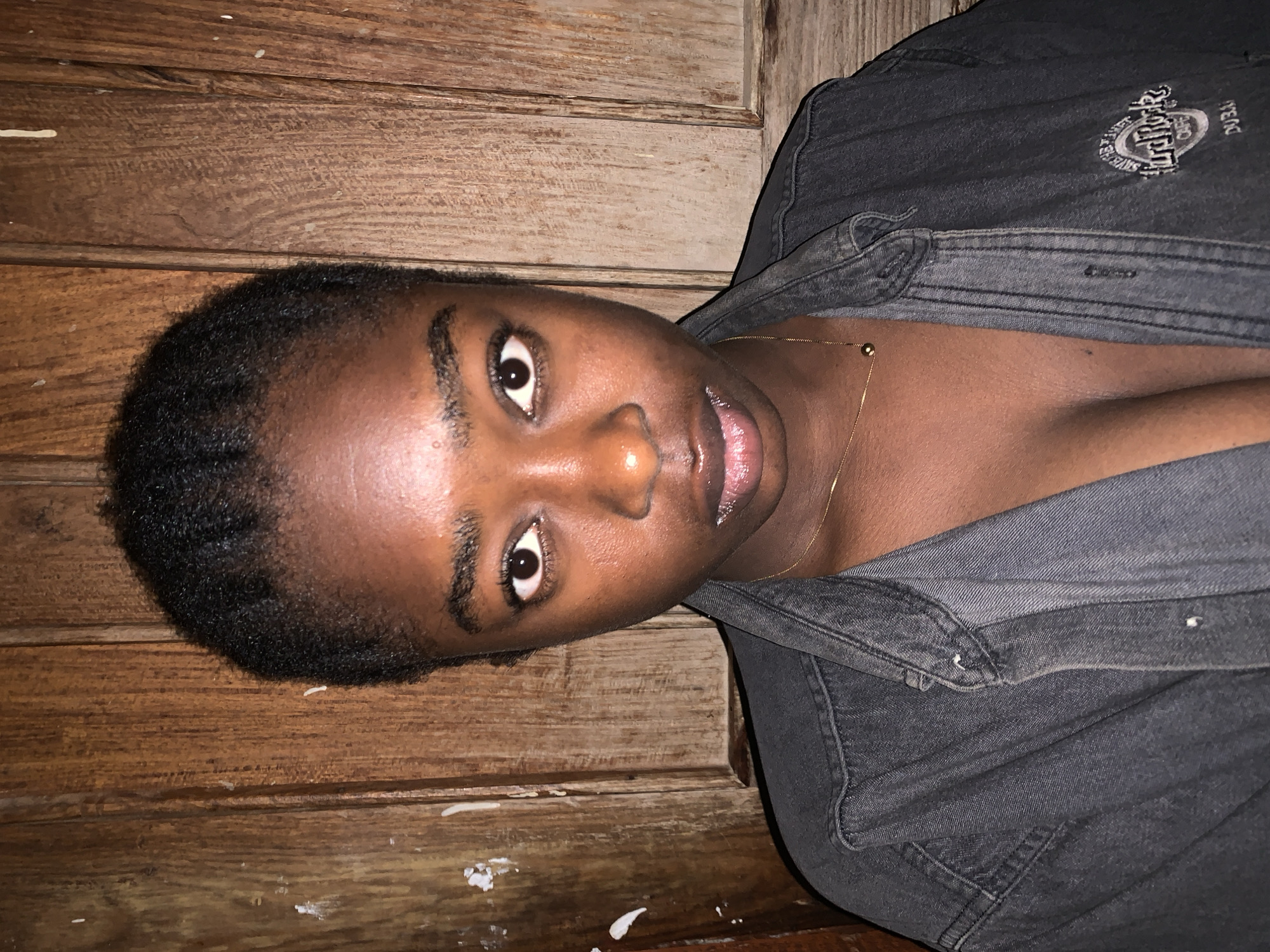 A person is seen indoors against a wooden wall, appearing to be lying on their side or leaning back. The individual has dark skin, dark, closely cropped hair, and is wearing a dark grey denim jacket. A thin gold necklace is visible on their neck. Part of the logo of the Hard Rock Cafe is visible on the jacket. The location is not detectable from the visual information. The image does not provide information for a forensic description beyond the person's appearance and the setting.