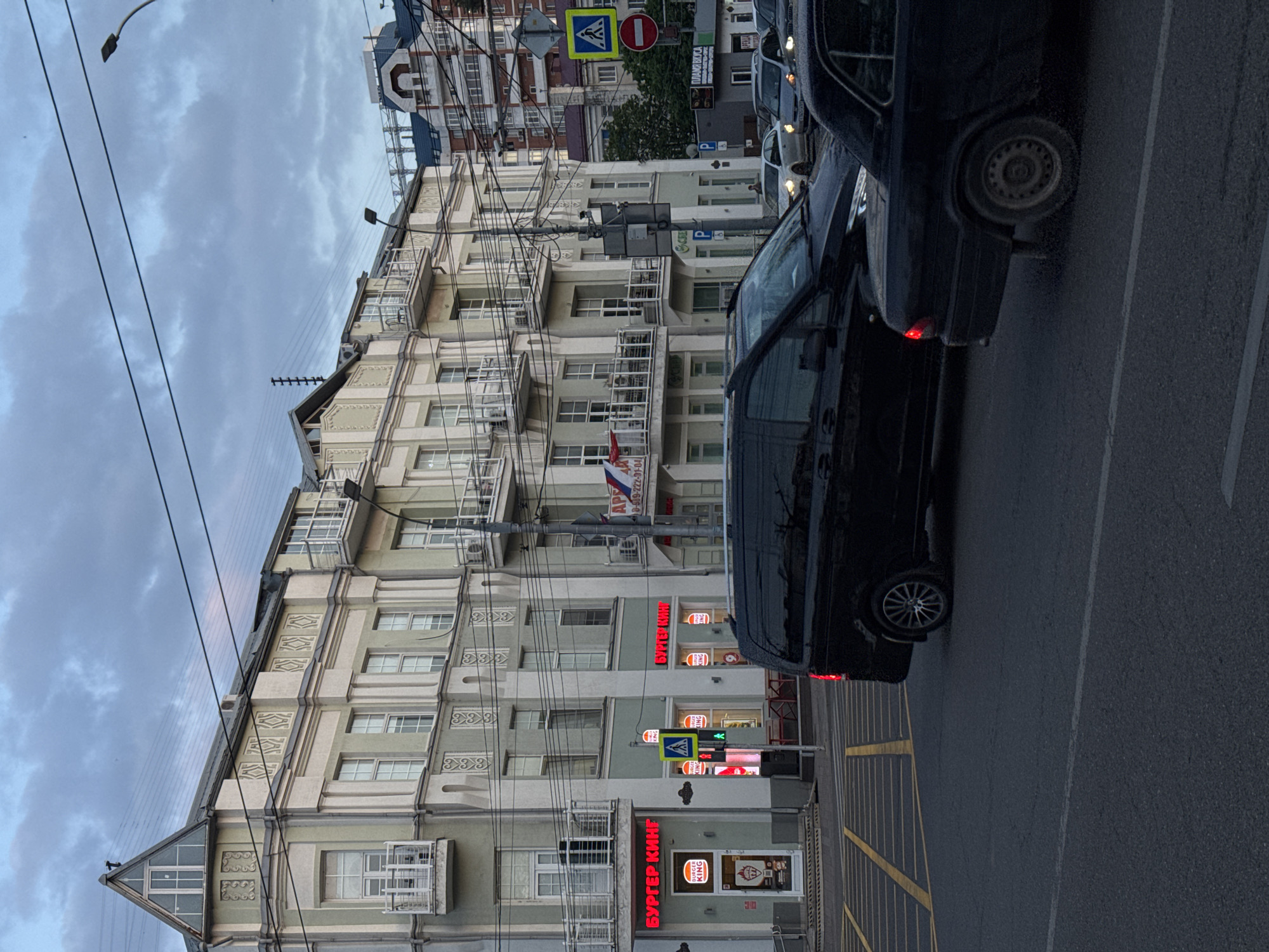 An urban street scene is captured, featuring a row of multi-story buildings under an overcast sky. The buildings have a classical architectural style with decorative elements and numerous balconies, and are painted in light shades of green and beige. Many power lines crisscross overhead.
Several dark-colored cars are visible, parked or moving along the street to the right of the frame. One car's rear red taillight is illuminated. The asphalt road shows painted yellow parking lines.
No people are clearly visible. The time of day appears to be daylight, possibly late afternoon or an overcast day, given the diffused light.
Notable details include various street signs, such as a pedestrian crossing sign and a red "no entry" sign. Red neon signs stand out against the buildings. Visible text includes "БУРГЕР КИНГ" (Burger King) displayed prominently in red on two signs, and "BURGER KING" and "KING" on the building facade. Other partial text includes "ПЛАМЯ ВКУСА" (Flame of Taste), "AP", a phone number "8-569-222-01-04", and isolated letters like "SCHE", "P", and "R". A small Russian flag is also visible on one of the buildings.