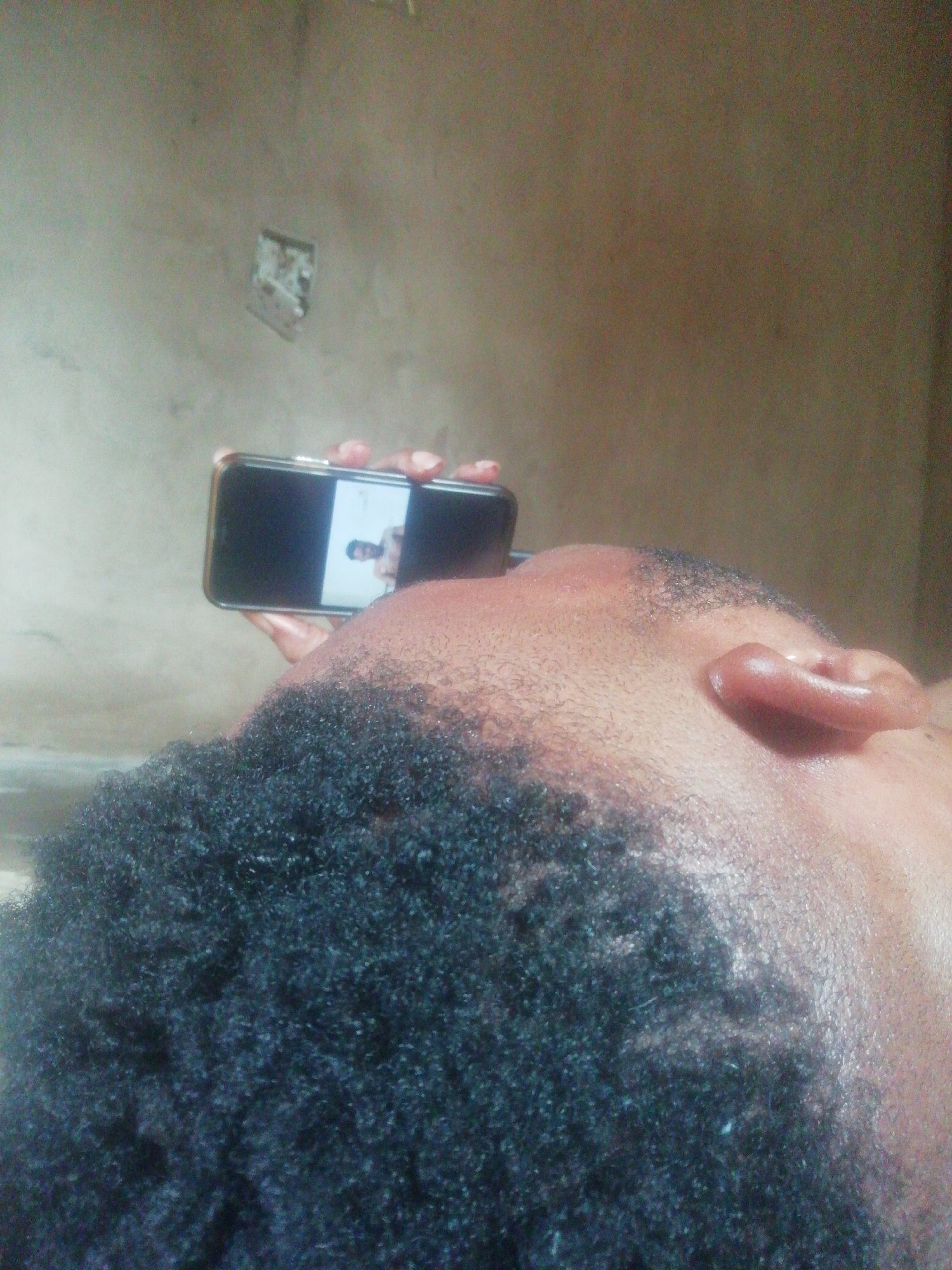 Here's a detailed description of the image:
**Scene:** The image appears to be a close-up perspective shot of a person lying down, viewed from above. The subject's head and shoulders are visible, filling much of the frame. The person is holding a smartphone, displaying an image of another individual on the screen.
**People:**
* **Subject:** An individual is seen from a close perspective, with their face partially obscured. The person has dark skin and short, curly black hair. The subject is lying down, facing the camera.
* **Image on Phone:** The smartphone displays an image of another person with dark hair and fair skin.
**Objects:**
* **Smartphone:** The subject is holding a black smartphone, displaying an image.
* **Wall:** A plain wall is visible in the background, with a damaged patch.
**Actions and Interactions:**
* The subject is holding a smartphone and viewing an image of another person.
**Environment:**
* The environment is indoor, possibly a room with a plain wall.
**Location:** The city and state are listed as "Unknown, Unknown."