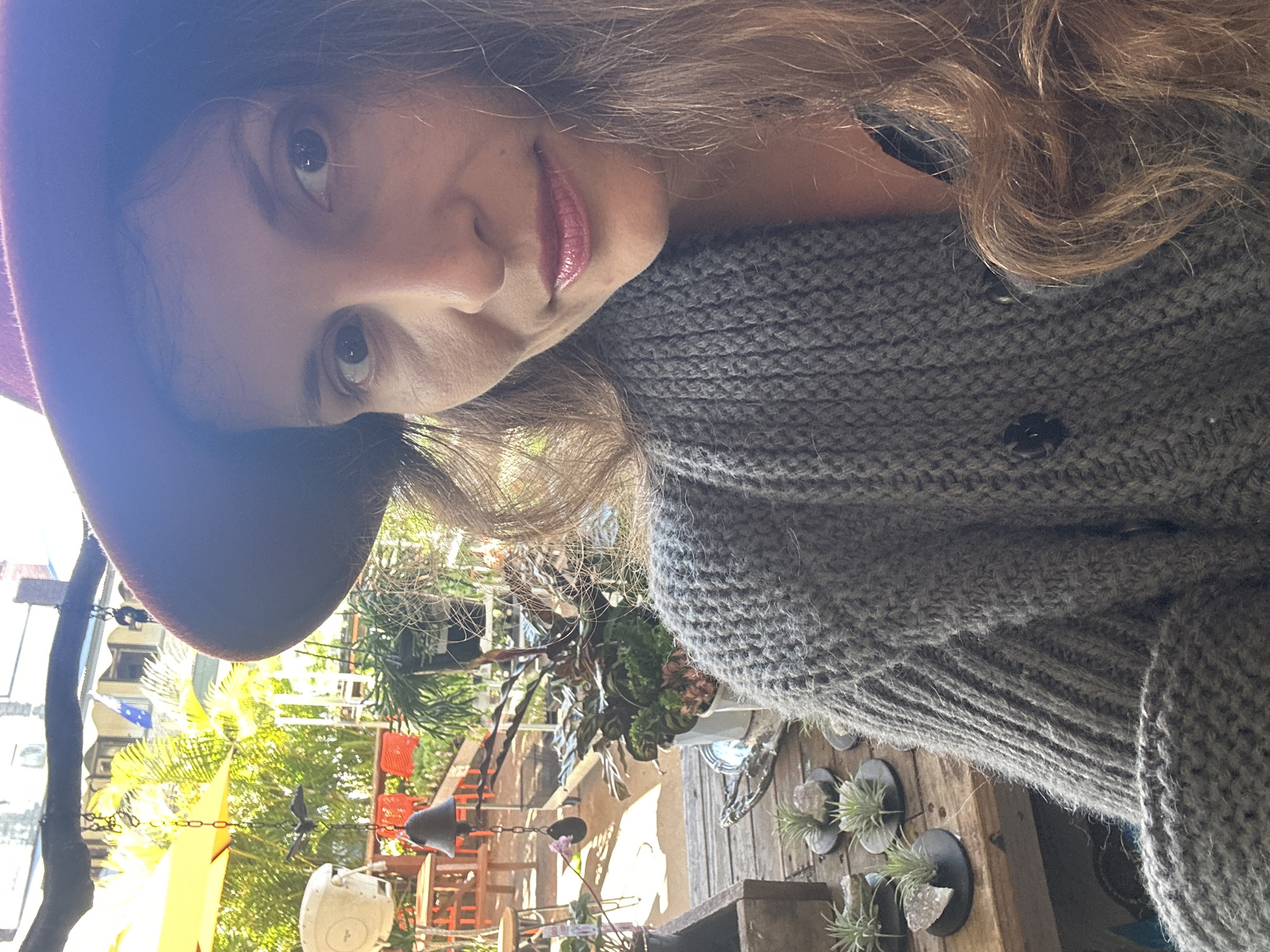 The image is a close-up, slightly angled selfie of a woman wearing a burgundy hat and a grey knitted cardigan. She has long brown hair and is looking towards the camera with a neutral expression. The background appears to be an outdoor patio or garden area, with plants, tables, chairs, and a yellow umbrella visible. Sunlight is casting shadows and highlights throughout the scene. There are no discernible landmarks that would place this location specifically in Bellingen, Australia.
