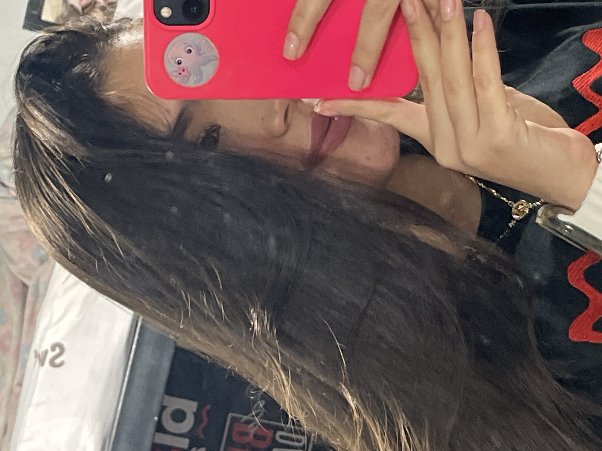 A young woman takes a mirror selfie in Juan Griego, Venezuela, holding a bright pink smartphone in her right hand, obscuring part of her face. Her long, dark hair falls over her face and shoulder. She is wearing a black shirt with a red wavy design. A necklace with a gold pendant is visible around her neck. Her fingernails are painted a light pink. A circular sticker of a cartoon octopus is on the back of her phone. The background is blurred but appears to be an indoor setting with some bedding and a shelf with items.