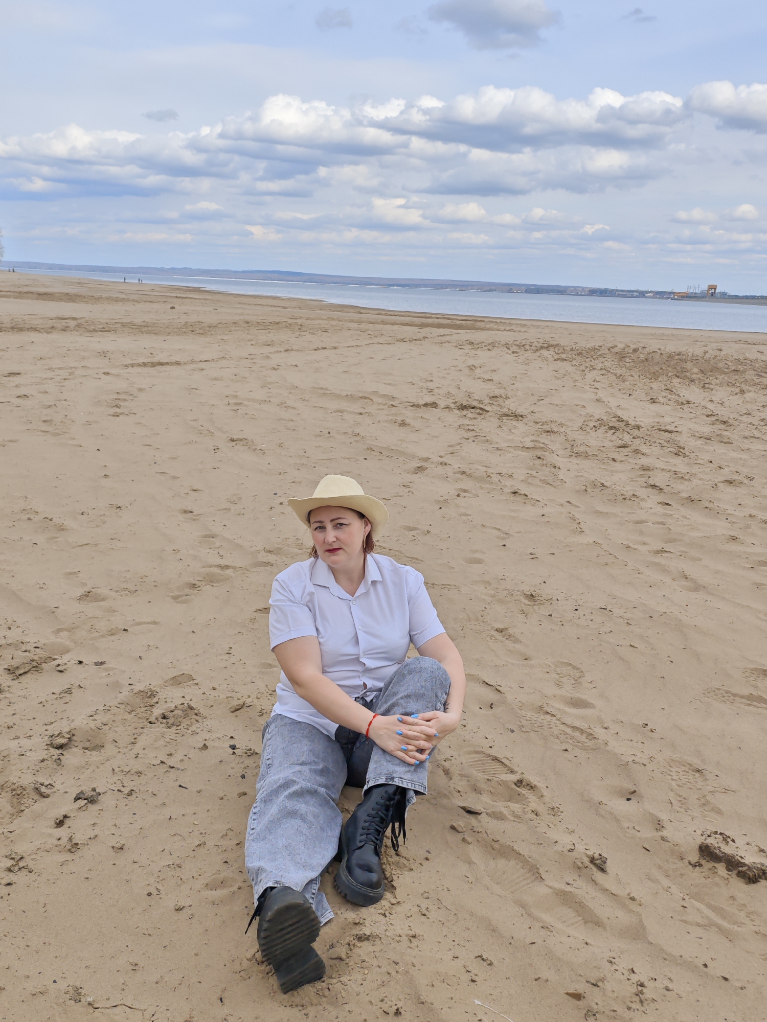 A woman is seated on a sandy beach. She wears a light-colored short-sleeved shirt, gray jeans, and black boots. A straw hat is on her head. She is hugging her legs with both hands. The beach extends into the distance towards a body of water. On the opposite side of the water is a city. The sky is cloudy. The location is Noviy Urengoy, Russia.