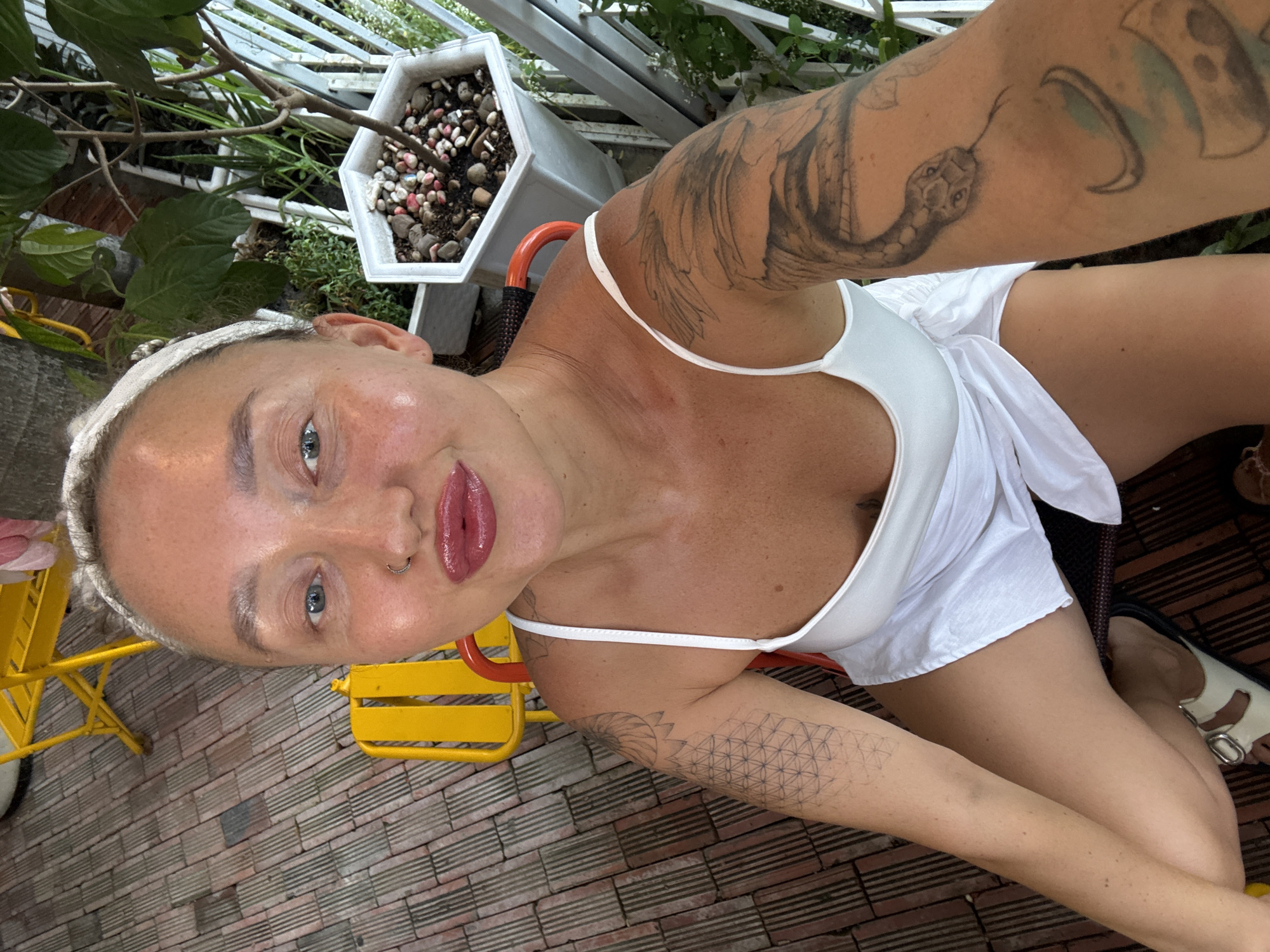 A woman is sitting on a yellow chair outdoors, with a brick-like patio beneath her and a balcony railing with plants in the background. She has blonde hair pulled back and is wearing a white dress. She has visible tattoos on her arms, including a snake on her right arm. She is looking towards the camera and appears to be smiling slightly. The scene is set in Nha Trang Ward, Vietnam, suggested by the specific type of brick and foliage common in the region.