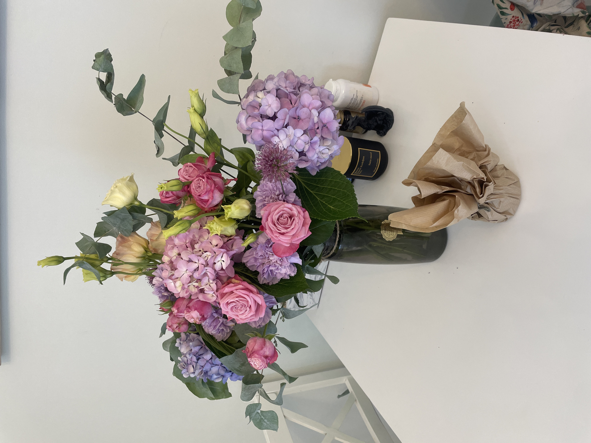 A floral arrangement of hydrangeas, roses, lisianthus, and eucalyptus is displayed in a dark glass vase on a white table. To the right of the vase are a black candle in a black jar, a small black figurine, and two white bottles. A white chair is visible to the left of the table. The scene appears to be indoors, with a white wall in the background. The location is not specified.