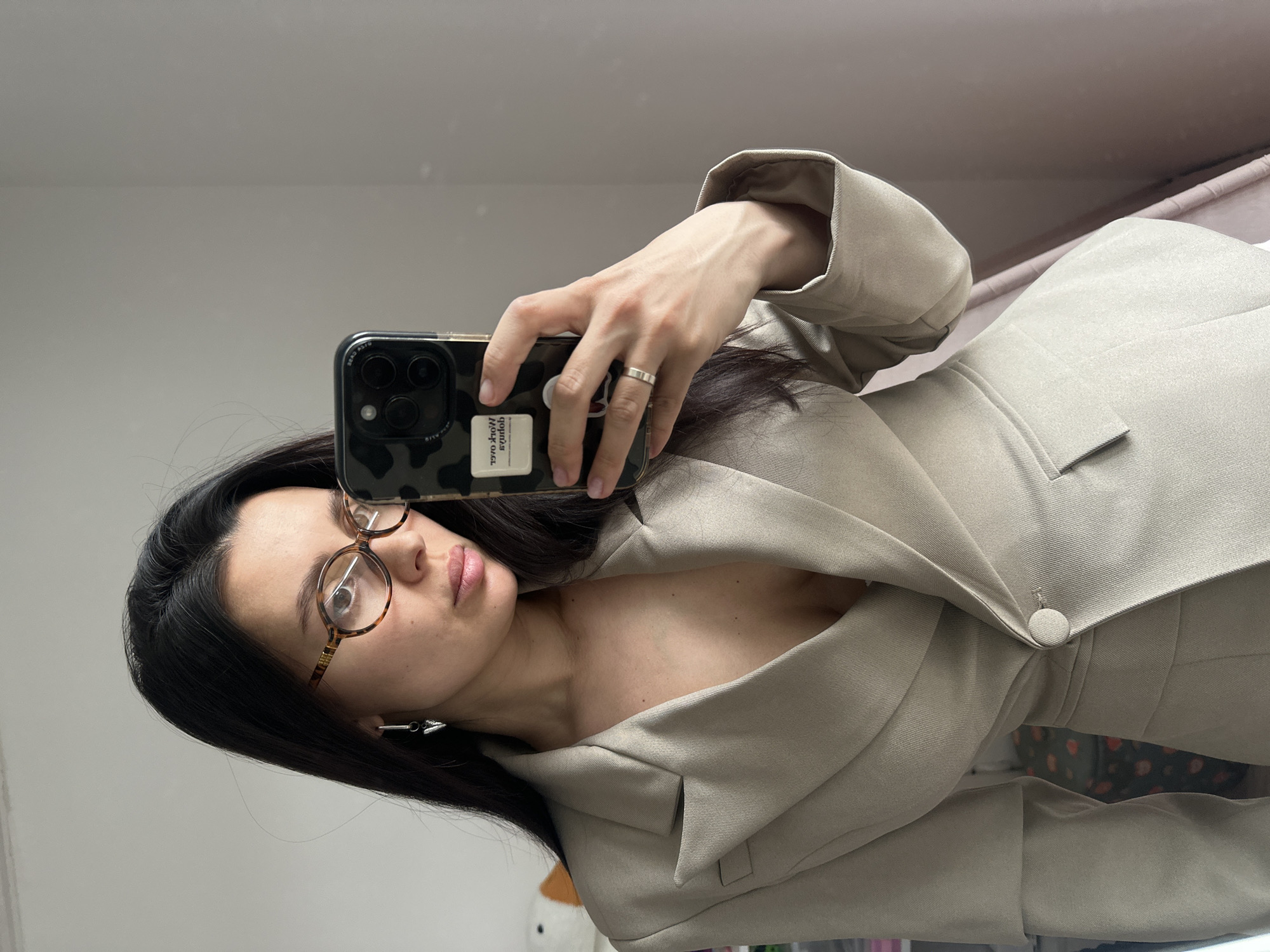 A woman is taking a selfie in what appears to be an indoor environment, possibly a home or apartment. She is holding a smartphone with a patterned case in her right hand. She is wearing glasses with a tortoise-shell frame and a light-colored blazer or jacket, which is buttoned. The background is mostly plain, with what seems like a wall. It is impossible to identify the city of Chaikovsky, Russia, from the provided image.