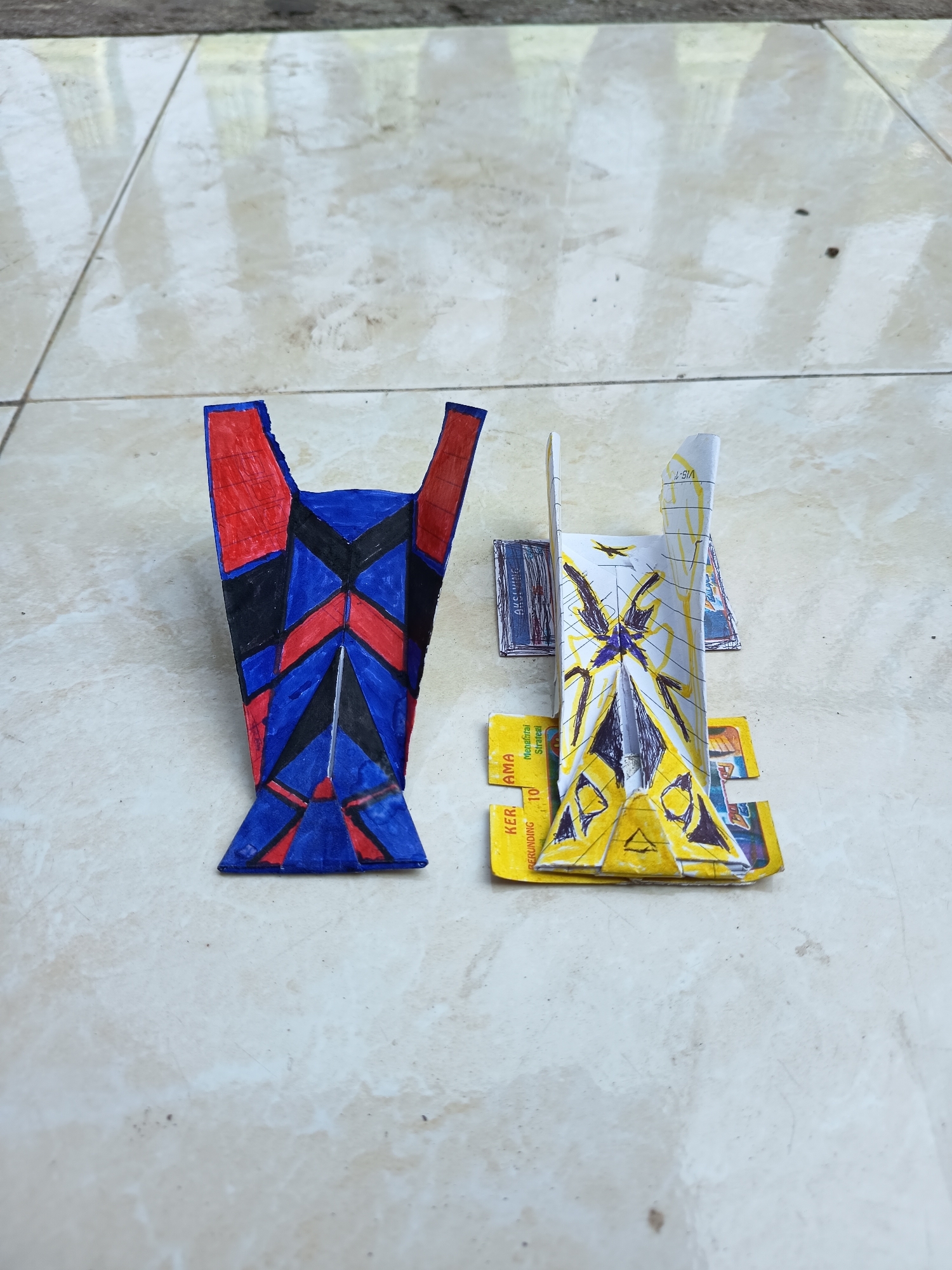 Two handmade paper toys, resembling futuristic vehicles or robots, are placed on a light-colored tiled floor. The toy on the left is primarily blue and red, with black accents forming a geometric pattern. It has a wide base that tapers upwards into two pointed sections. The toy on the right is predominantly yellow and white, with black markings and a purple star-like shape at its center. It has a more intricate, layered construction and appears to be made from folded paper, possibly incorporating pieces of packaging or cards. Small pieces of printed material with text are visible underneath and around the yellow and white toy. The tiled floor shows a subtle pattern of light and shadow, reflecting light from an unseen source. The background is out of focus, but appears to be an outdoor setting with a hint of greenery and a textured surface. The image was taken in Gebangan, Indonesia.