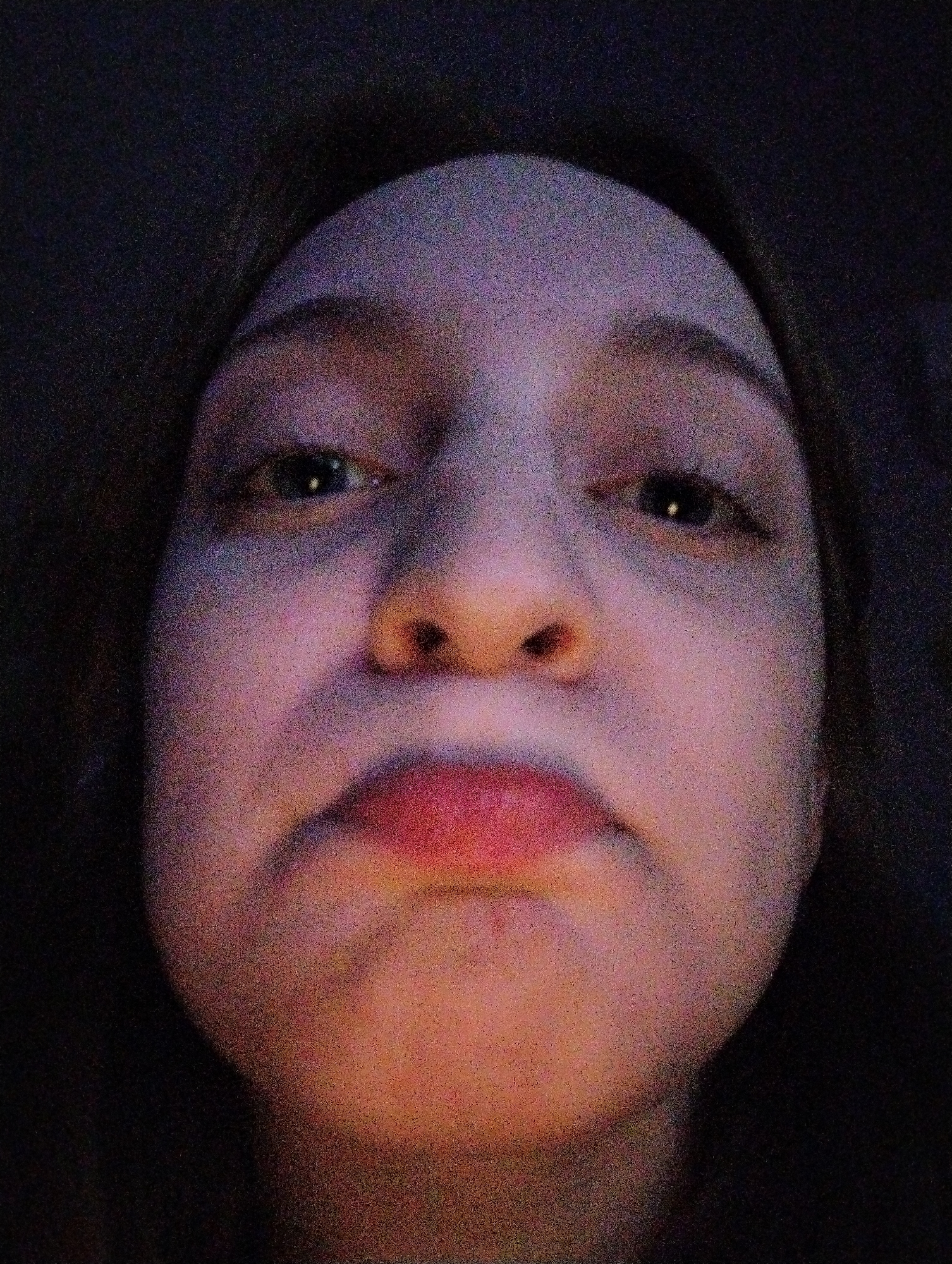 A close-up, poorly-lit, front-facing selfie of a person's face. The person appears to have fair skin, dark hair, and is looking directly at the camera. The lighting is uneven with a light source below the face, illuminating the chin and lips. The background is dark and indistinct. There is no visible indication of Saratov, Russia, or any other specific location. No specific actions, interactions, or objects are discernible beyond the person's face.