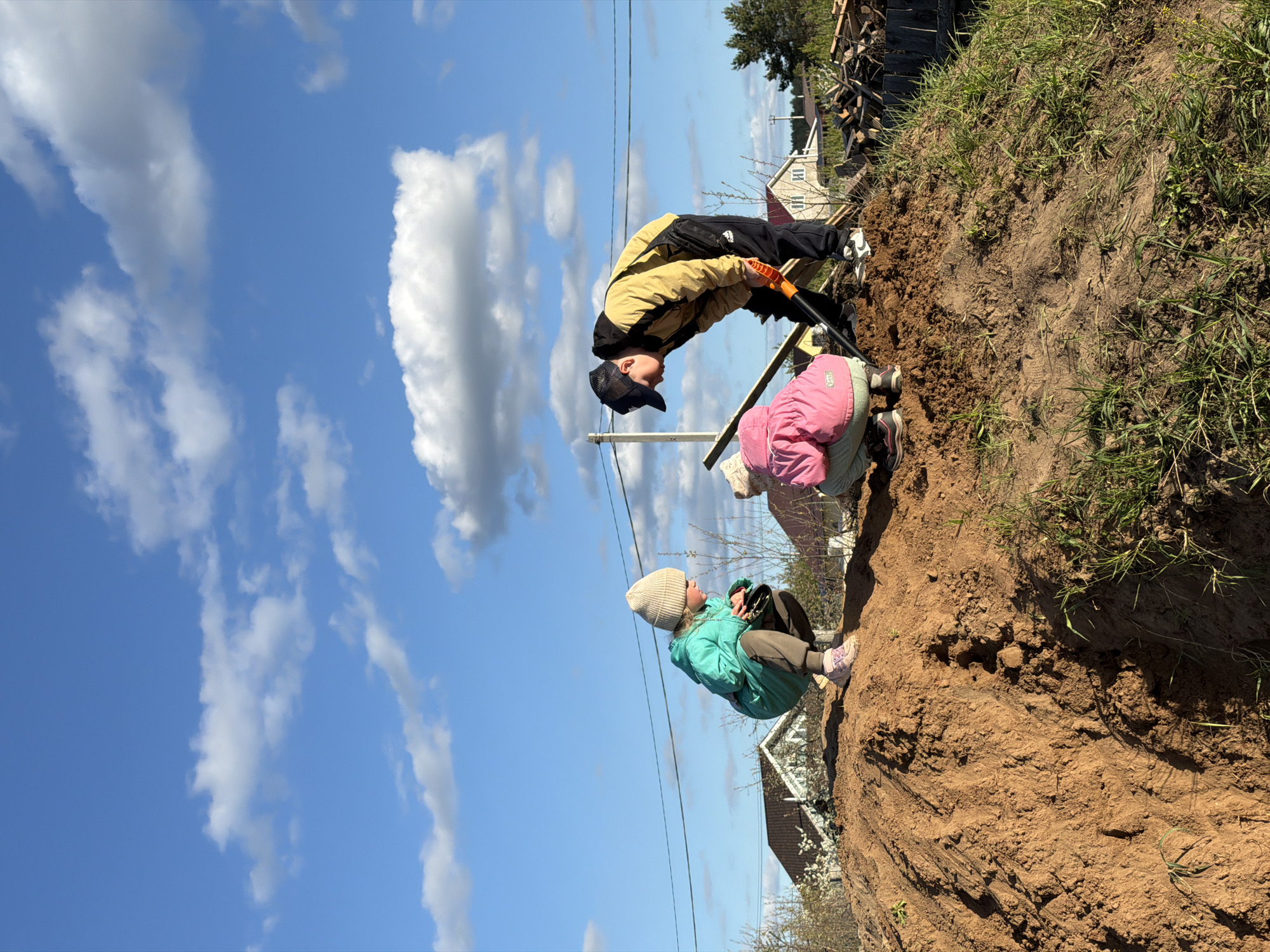 The image shows a man and two children digging into a dirt bank. The man, wearing a tan jacket and black pants, is using a shovel. One child, in a pink jacket, is crouched near the man. The other child, wearing a green jacket and a beige hat, is seated further along the dirt bank. The sky is blue with puffy white clouds. Power lines are visible. In the background, there are houses and trees. Given the request for a location in Chaikovsky, Russia, and lacking other contextual information, it is presumed that this image represents an outdoor activity in a residential area of Chaikovsky, Russia, where the individuals are excavating soil.