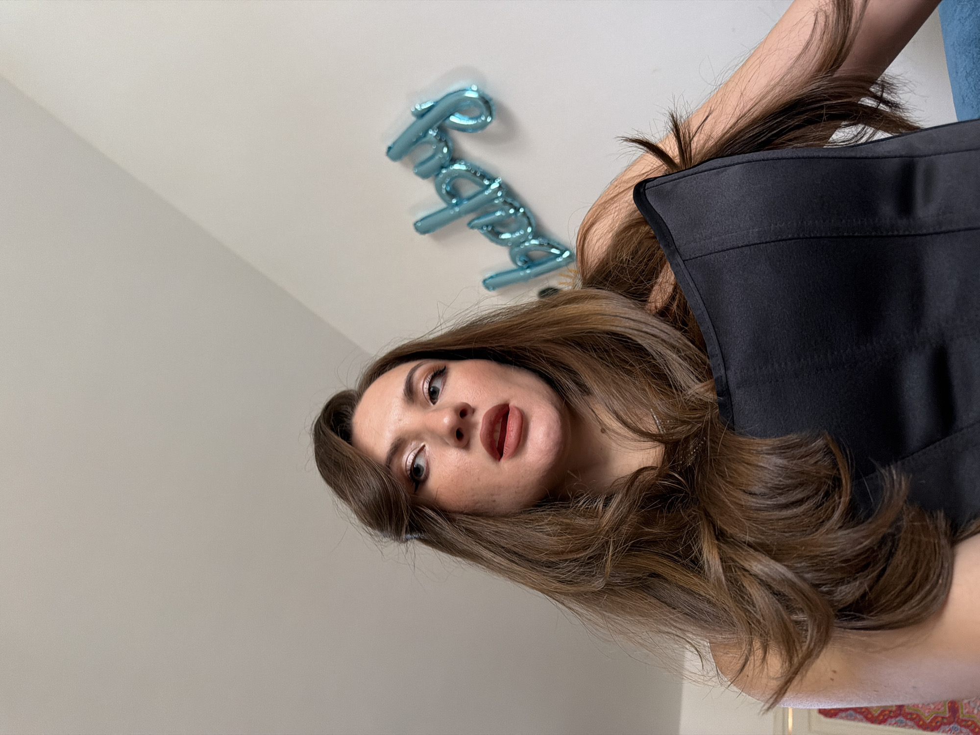 A low-angle, medium shot captures a woman with long, brown hair and a black corset top against a plain wall. A blue, foil balloon spelling "baby" is attached to the wall behind her. The woman is looking up and to her left, with her mouth slightly open. The lighting is natural, casting soft shadows. The background is a light gray wall, and the floor is not visible. The image appears to be taken indoors, possibly in a residential setting, with no discernible features suggesting a specific location within Sharm El Sheikh, Egypt.
