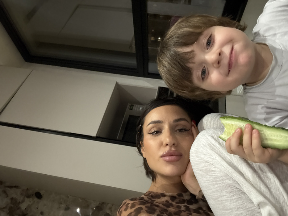 A woman with short dark hair and bold makeup, wearing a brown leopard print top, poses cheek-to-hand with a child. The child has light brown hair and is holding a partly eaten cucumber. The background features white kitchen cabinets, a black-framed window, and a microwave. The photo is taken at an angle, looking slightly upward.