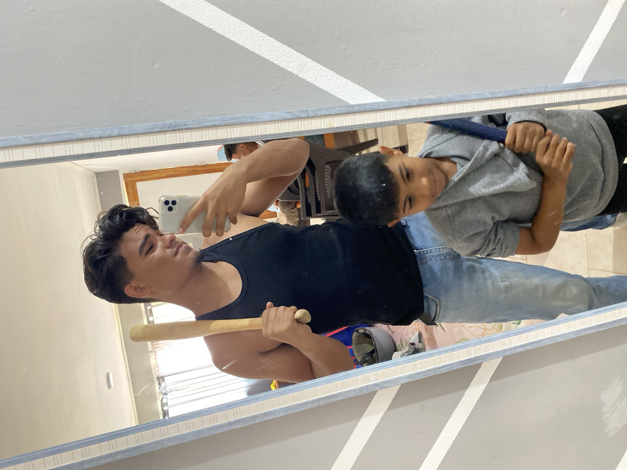 A mirror selfie shows a young man in jeans and a black tank top holding a baseball bat in one hand and a smartphone in the other. A younger boy wearing a grey hoodie and black pants stands next to him in the reflection, holding a blue object. The background shows a tiled floor and a glimpse of furniture. The location is presumably a residential interior. The city is Altagracia, Venezuela.