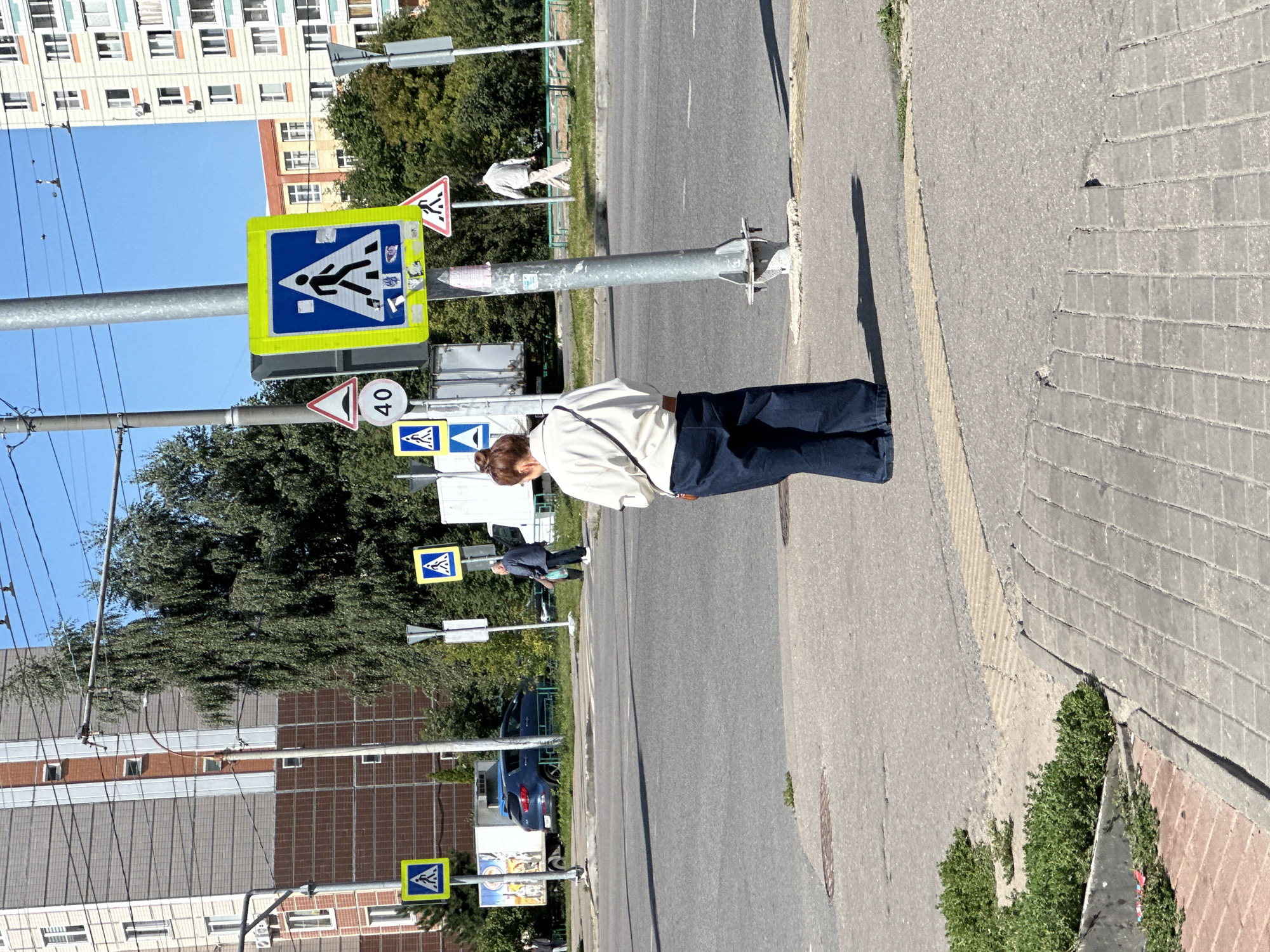 The image depicts a street scene in Podolsk, Russia, on a sunny day. A woman with brown hair pulled back in a bun stands on a sidewalk facing away from the camera, wearing a light-colored oversized shirt and dark blue wide-leg jeans. She is positioned near a metal lamppost.
In the background, across the street, there are several buildings, including a tall, light-colored apartment building and a red brick building. Trees and other vegetation line the street. Several traffic signs are visible, including a pedestrian crossing sign with a blue background and a white silhouette of a person walking, and a triangular sign with a red border and a white background depicting a person walking. A circular sign with "40" is also present.
On the street, there are vehicles, including a blue car, a white truck, and another white truck in the distance. Other pedestrians are visible in the background, one walking across the street and another walking on the sidewalk. The road surface is asphalt, and the sidewalk is paved with concrete slabs and cobblestones. The scene appears to be a typical urban intersection with pedestrian crossings.