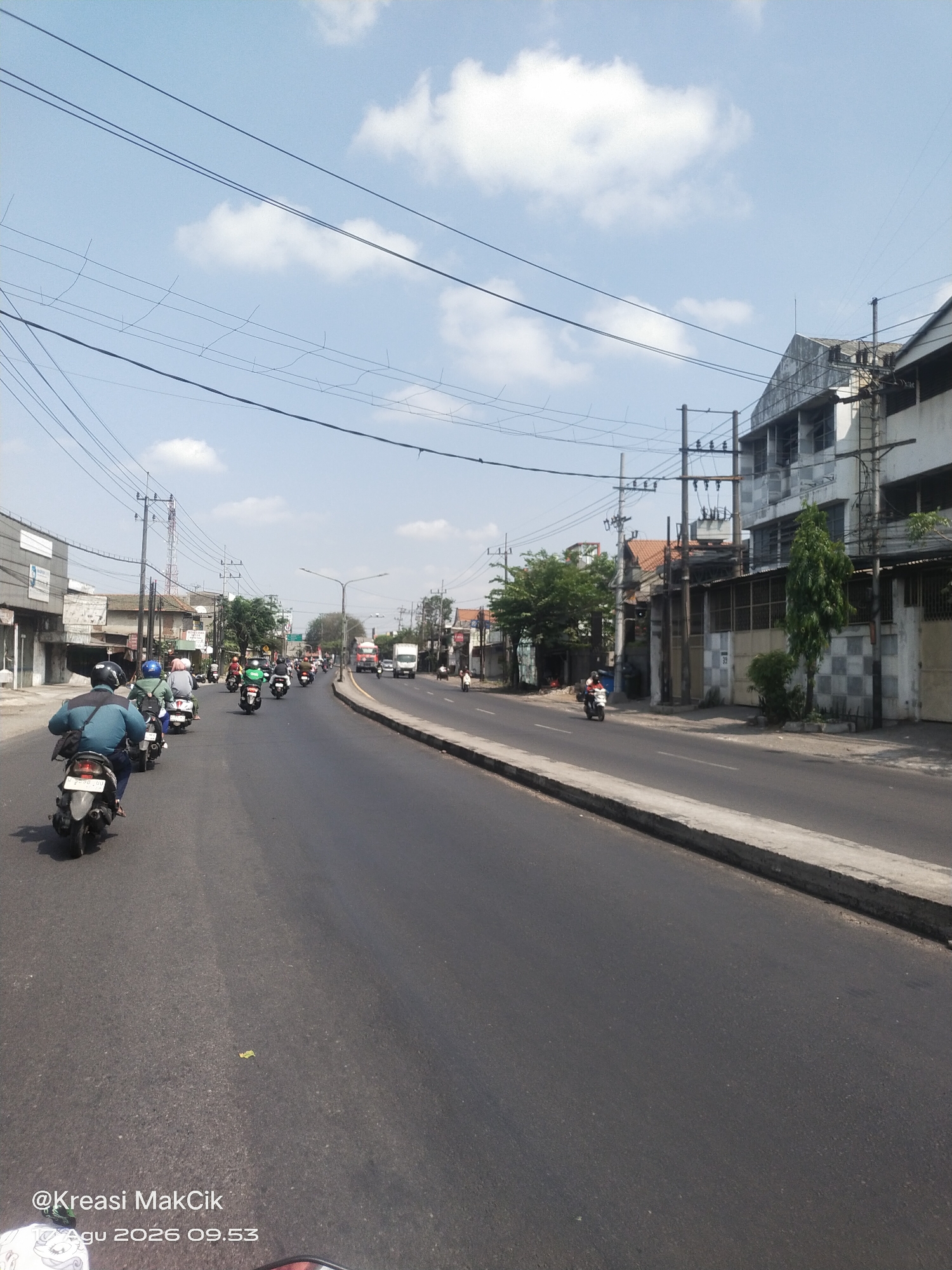 The image captures a daytime street scene in Kletek, Indonesia, characterized by a clear blue sky with scattered white clouds and overhead power lines. The perspective is from a moving vehicle, likely a motorcycle, looking forward along a paved road.
The road is moderately busy with traffic, primarily motorcycles, with several individuals riding them. Some motorcycles are in the foreground, while others are further down the road. A few cars and trucks are also visible in the distance, including a white truck and a smaller red truck.
On the right side of the road, there are buildings with a weathered, somewhat industrial appearance, featuring multiple stories and visible utility infrastructure like electrical poles and transformers. Trees are present along the roadside and in front of some buildings, with one tall, slender tree standing out near the right edge of the frame.
On the left side of the road, there are more commercial-looking buildings with varying facades, some appearing to be shops or businesses. A tall radio tower is visible in the mid-ground on the left.
The overall impression is of a functional urban environment with ongoing daily activities. The foreground shows the road surface and a portion of the vehicle from which the photo is taken, including a partial view of a helmet. A watermark or text overlay is visible in the lower left corner, reading "@Kreasi MakCik" and "Agu 2026 09.53", indicating the source and date of the image.