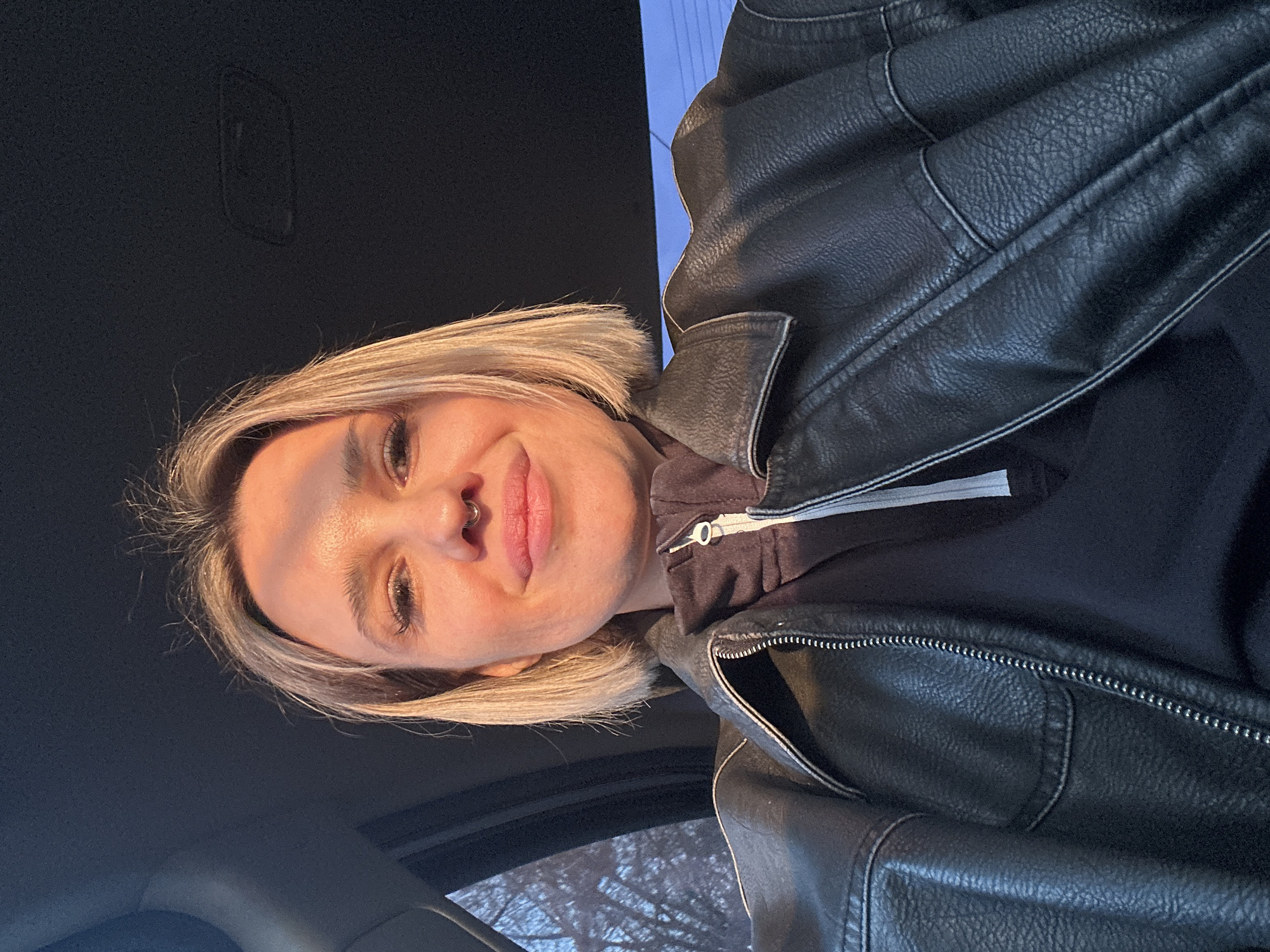 A female individual is depicted in a selfie-style photograph taken inside a vehicle in Rostov-on-Don, Russia. The subject, appearing to be in her mid-20s to early 30s, has blonde, chin-length hair and a silver-colored septum piercing. She is wearing a black leather-like jacket over a dark-colored, possibly black or dark grey, zippered hoodie or sweatshirt. Strong, warm sunlight illuminates the right side of her face and jacket, casting shadows and indicating direct sun exposure, likely from a side window. The subject is looking directly at the camera with a slight smile. Through the vehicle's left-side window, bare tree branches are visible against a light blue sky, suggesting an outdoor environment during a colder season or late afternoon. The vehicle's dark interior headliner is visible at the top of the frame.