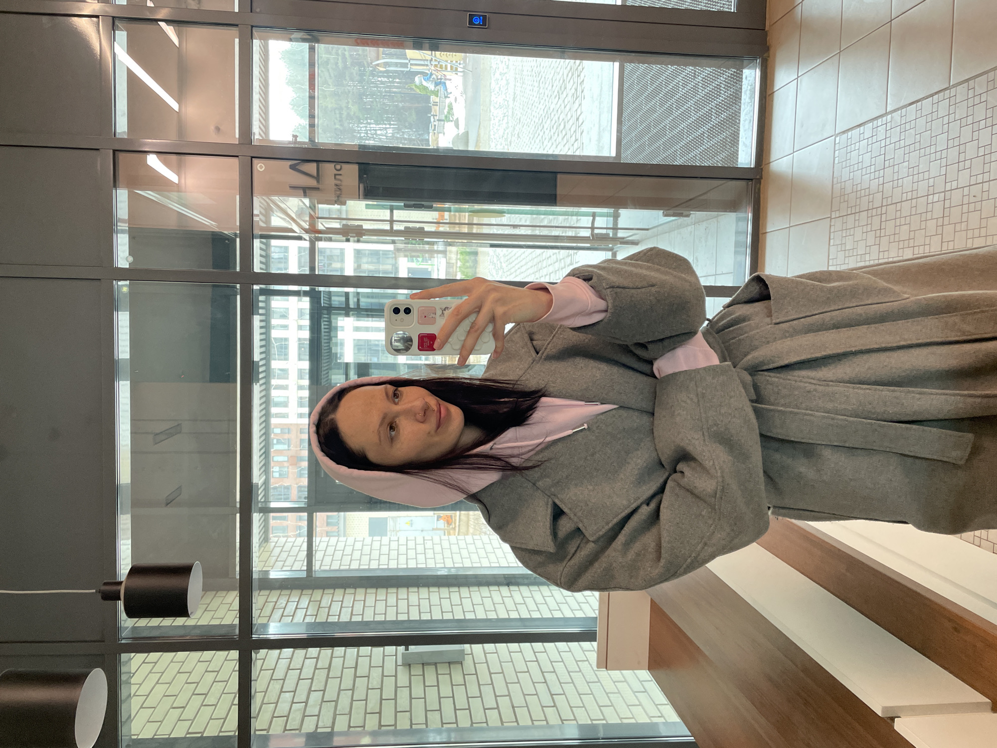 A woman takes a selfie in front of a window. She wears a grey coat over a pink hooded sweatshirt. The window looks out onto a street and buildings. The location is in Izhevsk, Russia.