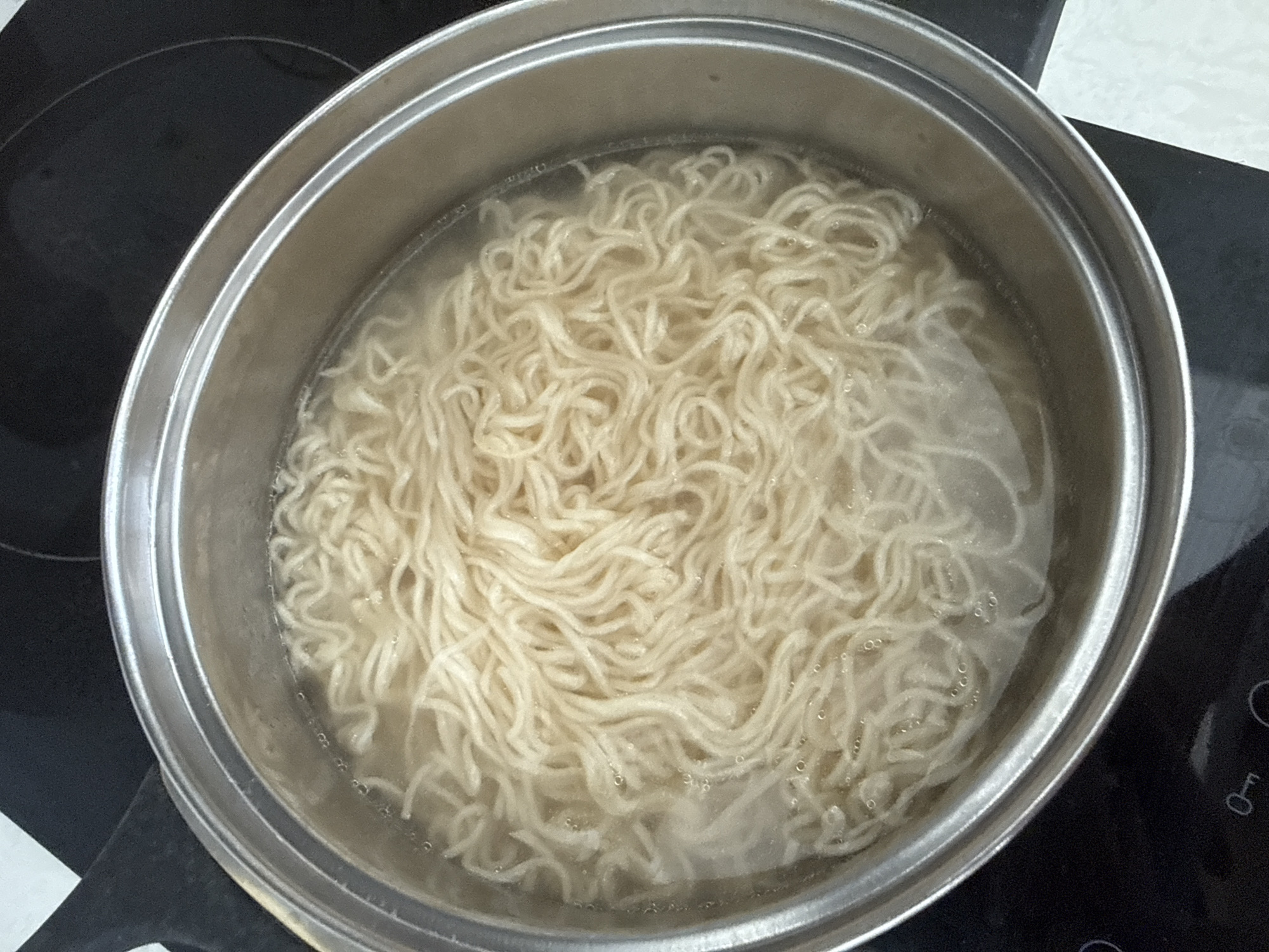A close-up, overhead shot shows a stainless steel pot filled with water and instant ramen noodles on a black stovetop. The noodles are tightly packed and submerged in clear water, with a few small bubbles forming on the surface, indicating the water is heating or boiling. The pot has a slightly raised rim and is placed on a modern electric stovetop. The stovetop has circular heating elements indicated by faint lines. The environment appears to be a kitchen in Burgas, Bulgaria, given the stovetop design which is common in European households. There are no people visible in the image.