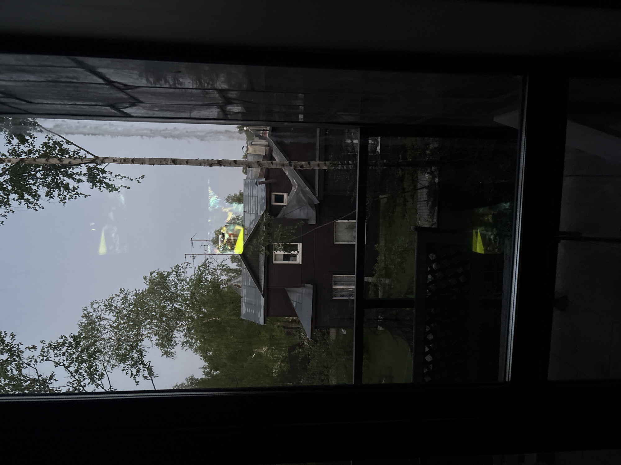 A view from a window, likely in a building, reveals a street scene in Saint Petersburg, Russia. The scene is dominated by residential houses with gray metal roofs and dark exteriors, possibly painted wood. A birch tree with bright green leaves stands to the left. The sky is overcast, suggesting either early morning, late evening, or a cloudy day. There is a possible television aerial on top of the houses. The image is partly obscured by the dark frame of the window through which the photo is taken. The scene looks like it might be in a somewhat rural or suburban area of Saint Petersburg.
