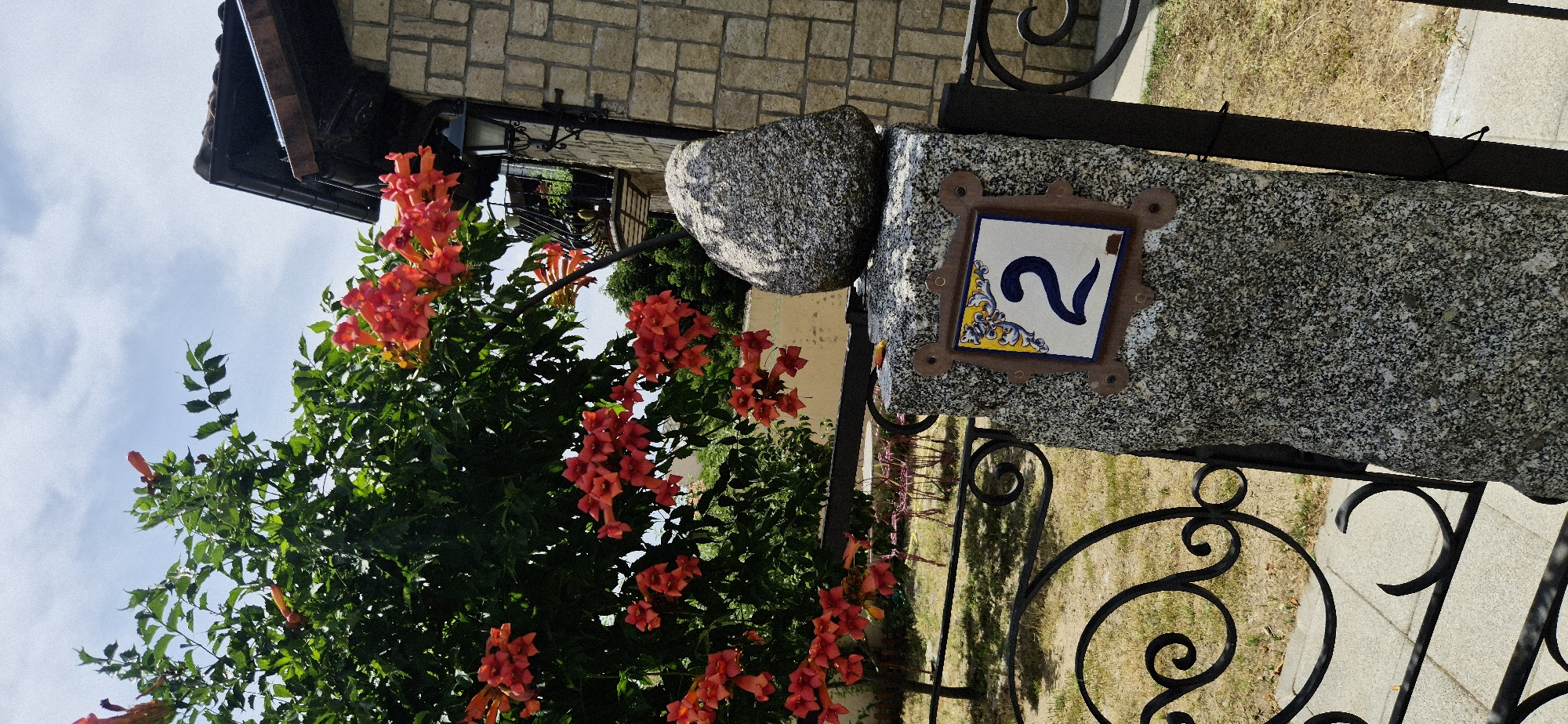 The image shows a close-up view of a decorative house number "2" mounted on a speckled granite pillar. The number is on a white tile with blue lettering and decorative yellow and blue accents around the border. The pillar is topped with a rough, gray rock and is part of a fence or gate. To the left of the pillar, a lush green bush with vibrant orange trumpet-shaped flowers is in bloom, with some flowers extending into the foreground. In the background, a stone-built house with a dark roof and a balcony with potted plants is visible. The scene is outdoors, with a bright sky and daylight present, suggesting a sunny day. The ground is covered with dry grass and a paved pathway. The overall impression is of a well-maintained property with natural beauty.