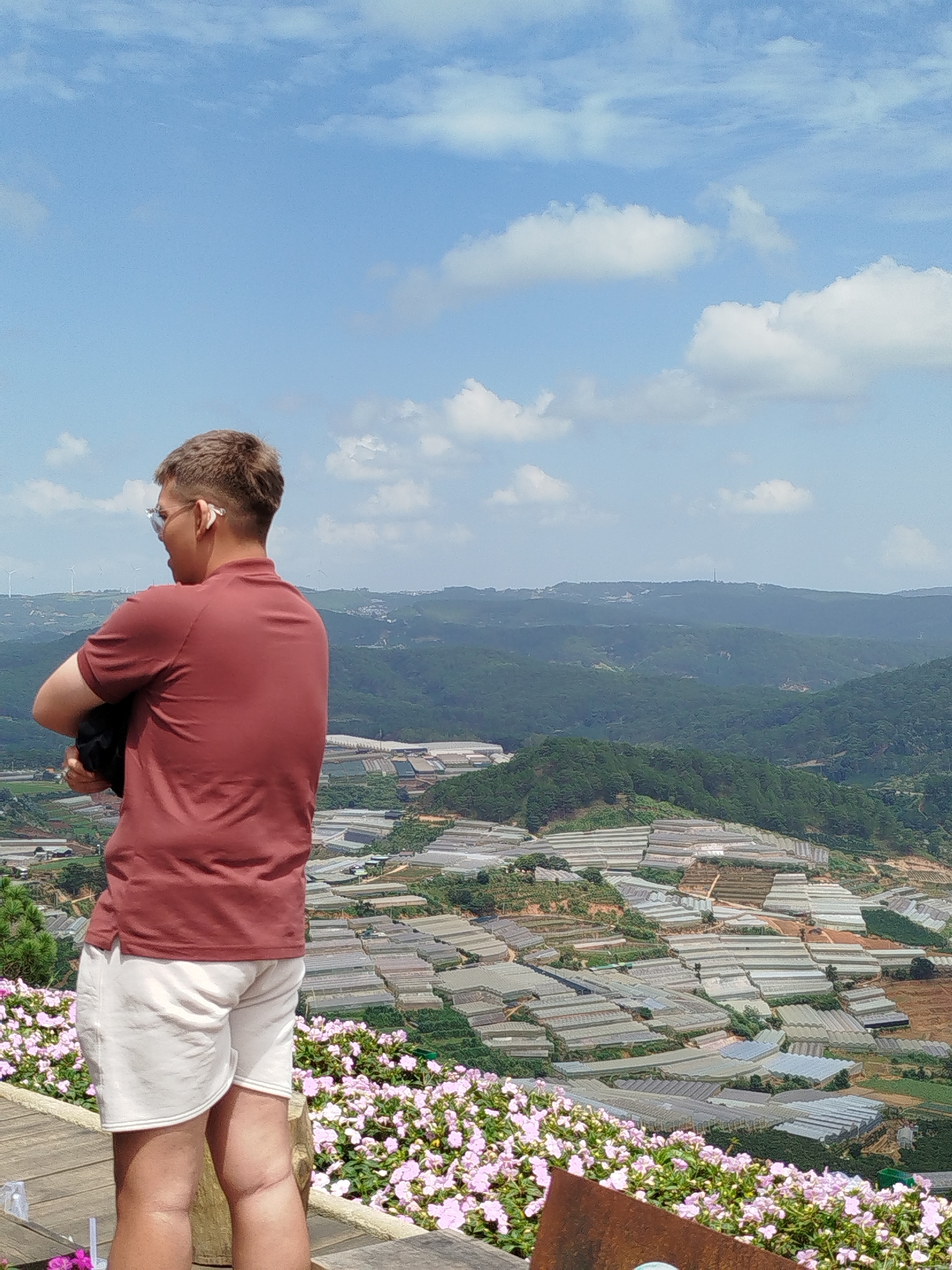 A person with short brown hair and sunglasses stands with their back to the camera, wearing a red polo shirt and white shorts. They are holding a dark object, possibly a bag, in their arms. The person is standing on a wooden platform, with a flowerbed of pink flowers in the foreground. Behind them, a vast landscape unfolds, characterized by rolling green hills dotted with numerous greenhouses, indicative of agricultural activity. In the distance, wind turbines are visible on the horizon, and the sky is a clear blue with scattered white clouds. The scene captures a view of the agricultural terraces and greenhouses commonly found in the Da Lat region, which is near Xuân Thành in Vietnam.