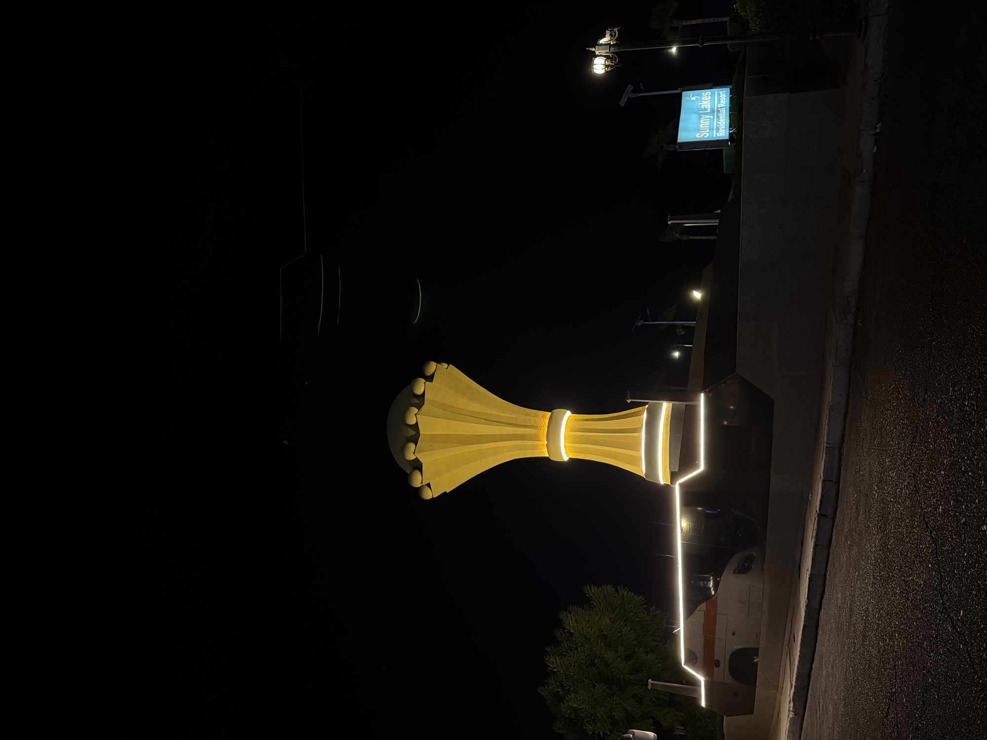 The image depicts a nighttime scene in Sharm El Sheikh, Egypt, featuring a distinctive yellow monument illuminated by white lights. The monument is tall and has a flared shape, with spherical elements at its apex. It stands on a concrete base that is also outlined with white lights. To the left of the monument, a tree with lush green foliage is visible. In front of the monument, a vehicle with an orange stripe is partially visible, parked behind a concrete barrier. A sign in the background reads "Sunny Lakes Residential Resort" in blue lettering against a white background. The foreground shows a paved road with a curb. The sky is dark, indicating it is nighttime, and streetlights are also visible.