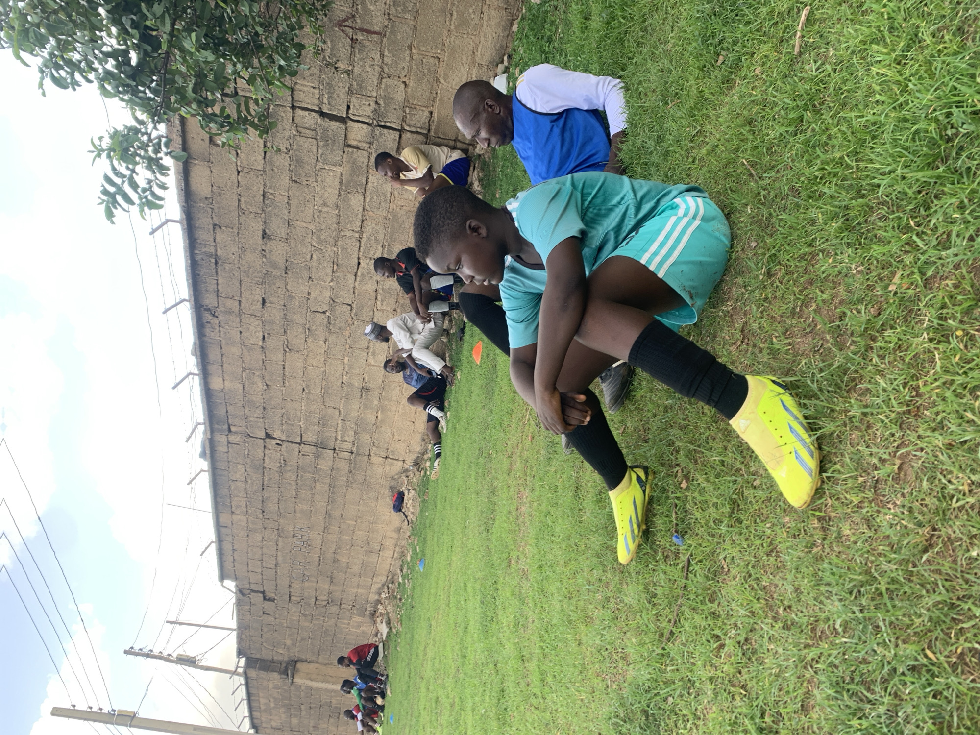A group of people are seated on a grassy field. The ground is covered with green grass, and there is a gray brick wall in the background. Overhead, there is an electrical line and a tree hanging over the wall. A young girl wearing a light blue athletic jersey and shorts, black socks, and yellow cleats is in the foreground. She is seated on the ground with her knees bent, leaning forward with her hands clasped together. Behind her, other people are also seated on the grass. One man is wearing a blue and white vest over a white long-sleeved shirt. In the background of the field are a few orange cones. The image was taken in Jos, Nigeria.