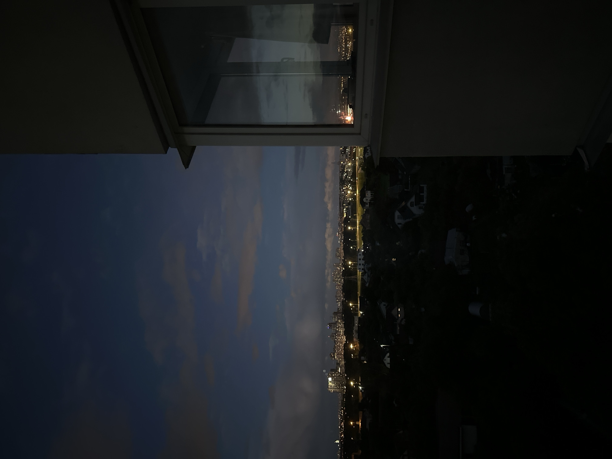 The image is a nighttime or twilight shot of a cityscape, likely from an elevated perspective, possibly a residential building. In the foreground, the right side of the frame is dominated by a portion of a building's exterior wall and a large window. The window reflects the twilight sky and distant city lights.
Beyond the immediate foreground, the scene unfolds into a panorama of urban and suburban areas. A dense cluster of buildings, including high-rises, forms the distant city skyline, illuminated by numerous lights. Closer to the viewer, a network of streets is visible, marked by streetlights. The middle ground features scattered houses and darker, densely vegetated areas, suggesting a transition from urban to more rural or suburban outskirts. The sky above is a deep blue, transitioning to darker tones at the top, with scattered clouds that are faintly illuminated from below by the city lights. The overall impression is one of urban sprawl at dusk or after dark. While the specific location is stated as Ждановіцкі сельскі Савет, Belarus, this visual information cannot be confirmed solely from the image itself. There are no people or animals visible in the frame.