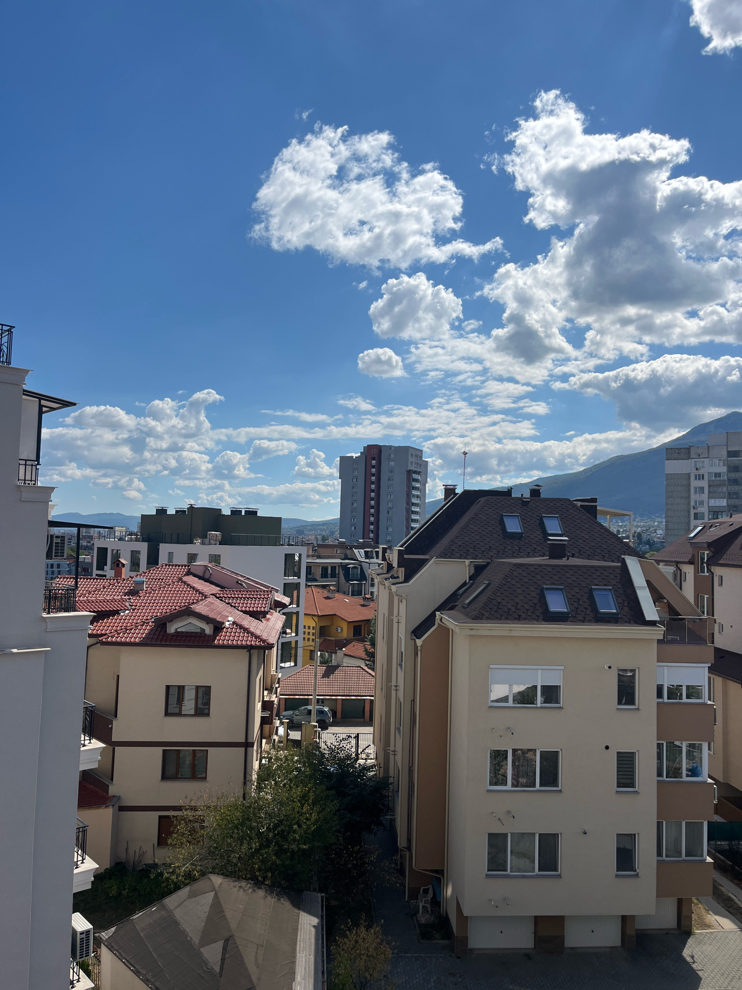 The image is a high-angle exterior shot of a residential area in Sofia, Bulgaria, on a clear day with scattered white clouds. The foreground features the white facade of a modern building with a balcony. Adjacent to it is a tan-colored building with a dark brown roof, several windows, and skylights. Further into the scene, a building with a red-tiled roof is visible, along with other residential structures of varying heights and architectural styles. In the background, taller apartment buildings and mountains are discernible against the blue sky. The overall impression is of a suburban or urban residential landscape. No people are visible in the image.