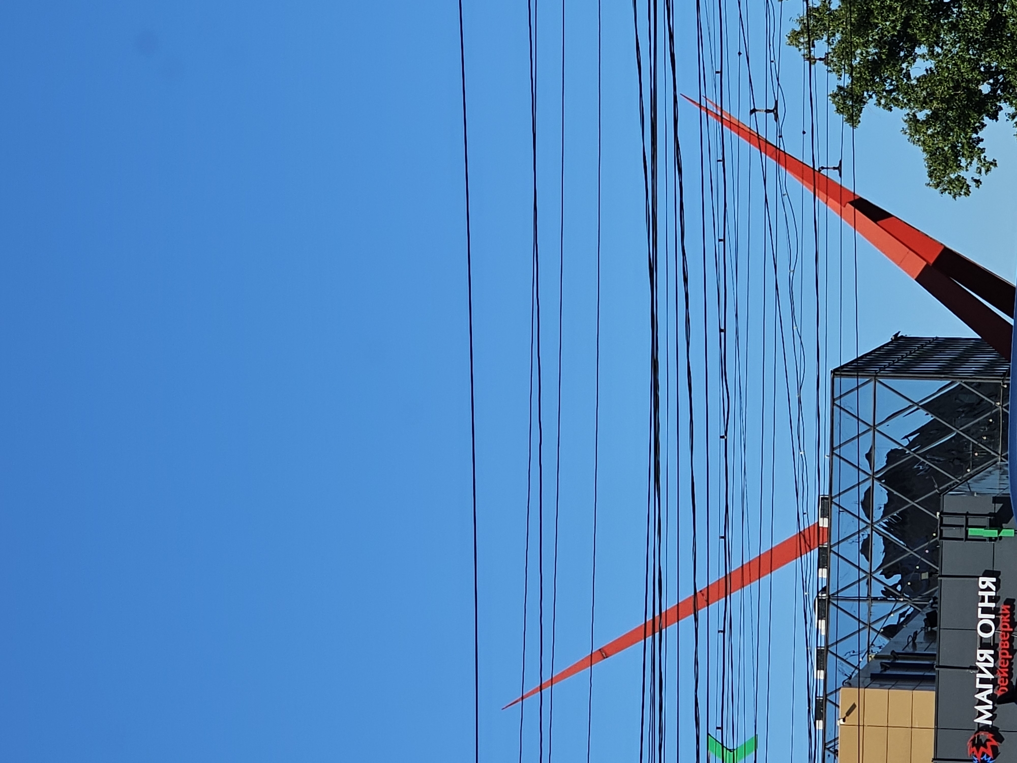 This is a photo taken in Nizhny Novgorod, Russia. The scene depicts a clear blue sky with a prominent network of black power lines stretching across the frame. Two bright red, sharp, pointed structures rise from behind a modern building. The building itself features a glass facade with a geometric pattern and a gray section with visible text. The text reads "МАГИЯ ОГНЯ" which translates to "Magic of Fire" and below it "фейерверки" meaning "fireworks". A green leafed tree is visible in the upper right corner. The lighting suggests it is daytime, likely sunny, given the clear sky and bright highlights on the building. The orientation of the photo is portrait mode, with the image appearing to be rotated 90 degrees counter-clockwise from its original capture. The overall mood is somewhat abstract due to the dominant sky and power lines, contrasted with the bold architectural elements. There are no people visible in the image.