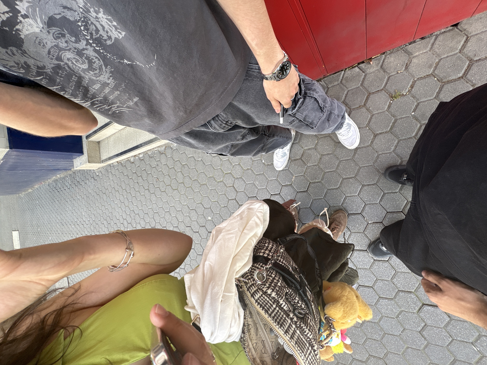 The image is a top-down view of a group of people standing on a hexagonal patterned grey pavement. A person on the left wears a light green top and a silver bracelet is visible on their left arm. Next to them is a pile of items, including a black and white checkered handbag, a white cloth, and a yellow stuffed animal. Another person wears a dark grey t-shirt and grey jeans, with a silver watch on their left wrist. They are holding a white object, possibly a pen or cigarette. Their feet are clad in white sneakers. Another person is positioned on the right side of the frame, dressed in black clothing and dark shoes. The edge of a red structure is visible in the upper right corner. The location cannot be determined from this image alone.