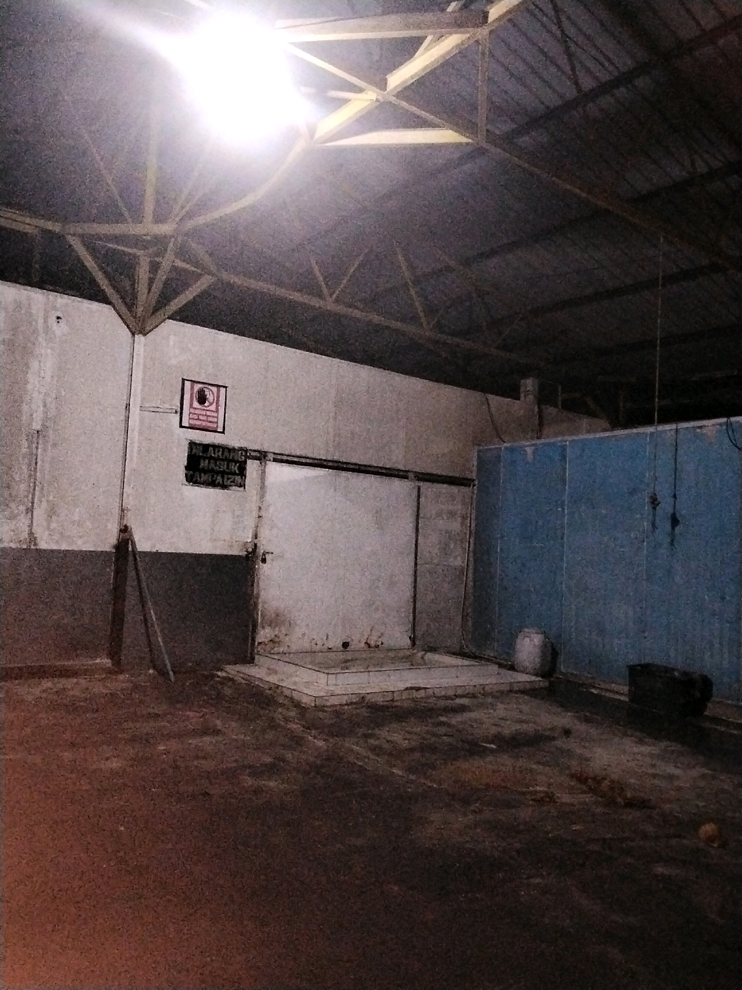 The image depicts an indoor, dimly lit space that appears to be an abandoned warehouse or industrial building located in Moro, Indonesia. The ceiling is constructed of metal sheeting supported by a triangular wooden truss system, with a bright overhead light fixture illuminating the left side of the scene. The walls are a dull, off-white color, stained and weathered, with a grey base near the floor.
On the left wall, a sign with a red border and a black symbol, possibly indicating prohibition, is visible. Below it, a black sign with white text reads "DILARANG MASUK TANPA IZIN," which translates to "NO ENTRY WITHOUT PERMISSION" in Indonesian. Adjacent to this sign is a large, off-white sliding door that appears to be closed or partially open. To the right of the door, there is a raised concrete platform with what seems to be a drain.
The right side of the image features a large, light blue wall. In front of this wall, on the floor, are several objects: a white cylindrical container, possibly a barrel, and a dark, rectangular object that could be a storage box or debris. The floor itself is uneven, covered in dirt, debris, and what appears to be spilled liquid or stains, giving it a dark, muddy appearance. In the far right of the image, a small, round, orange object rests on the floor. The overall atmosphere is one of disuse and neglect.