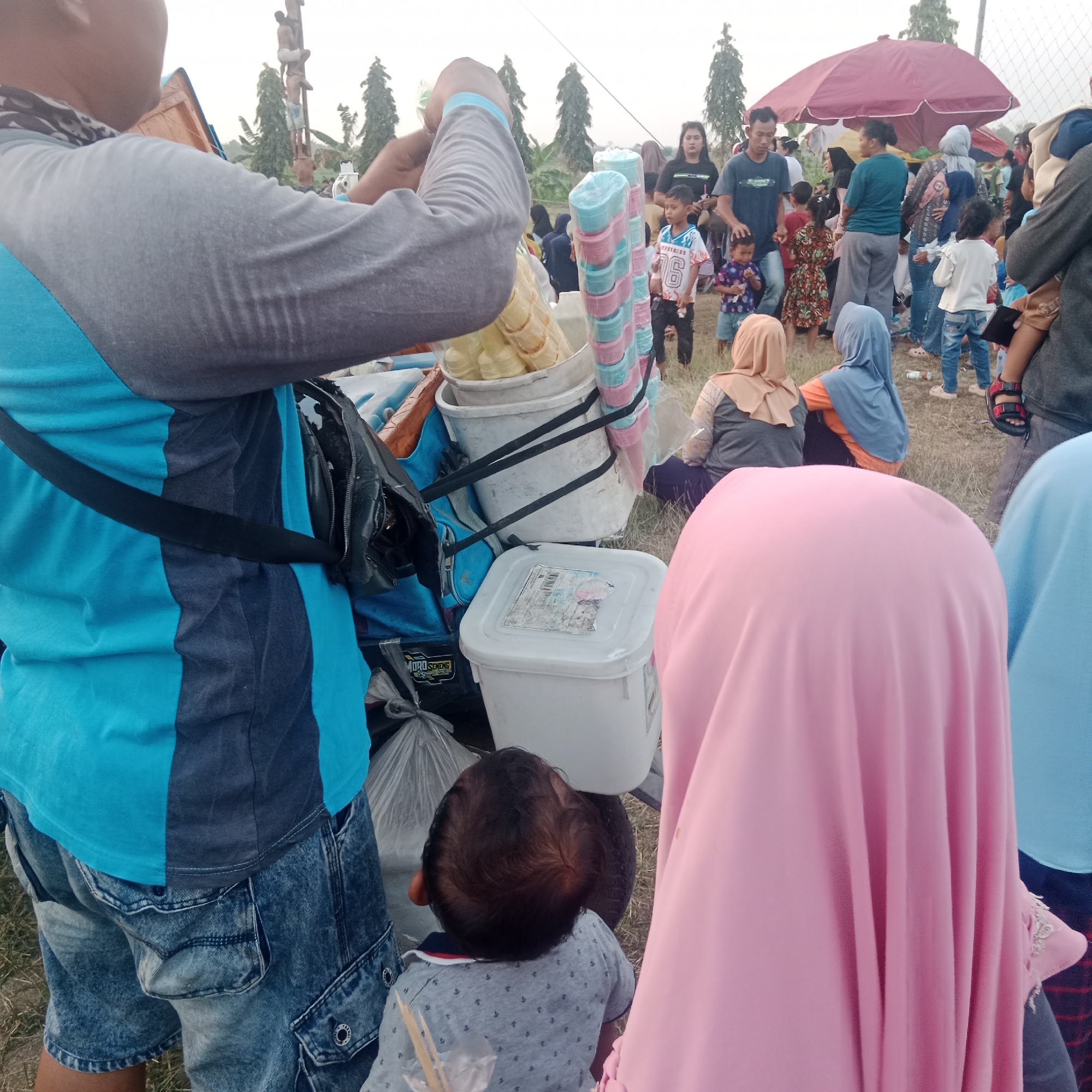 In Tegowanu Wetan, Indonesia, a crowd gathers outdoors, possibly for an event. The scene is dominated by a group of people, including adults and children, milling about in a grassy area. A vendor appears to be selling ice cream cones from a cart laden with various containers. In the foreground, a person in a blue and gray shirt and denim shorts is partially visible, and a child in a gray shirt with a patterned design is also present. A large pink hijab is prominent in the right foreground, obscuring the face of the wearer. In the background, a red umbrella provides shade, and several trees are scattered across the landscape. The lighting suggests it is daytime, likely late afternoon or early evening.