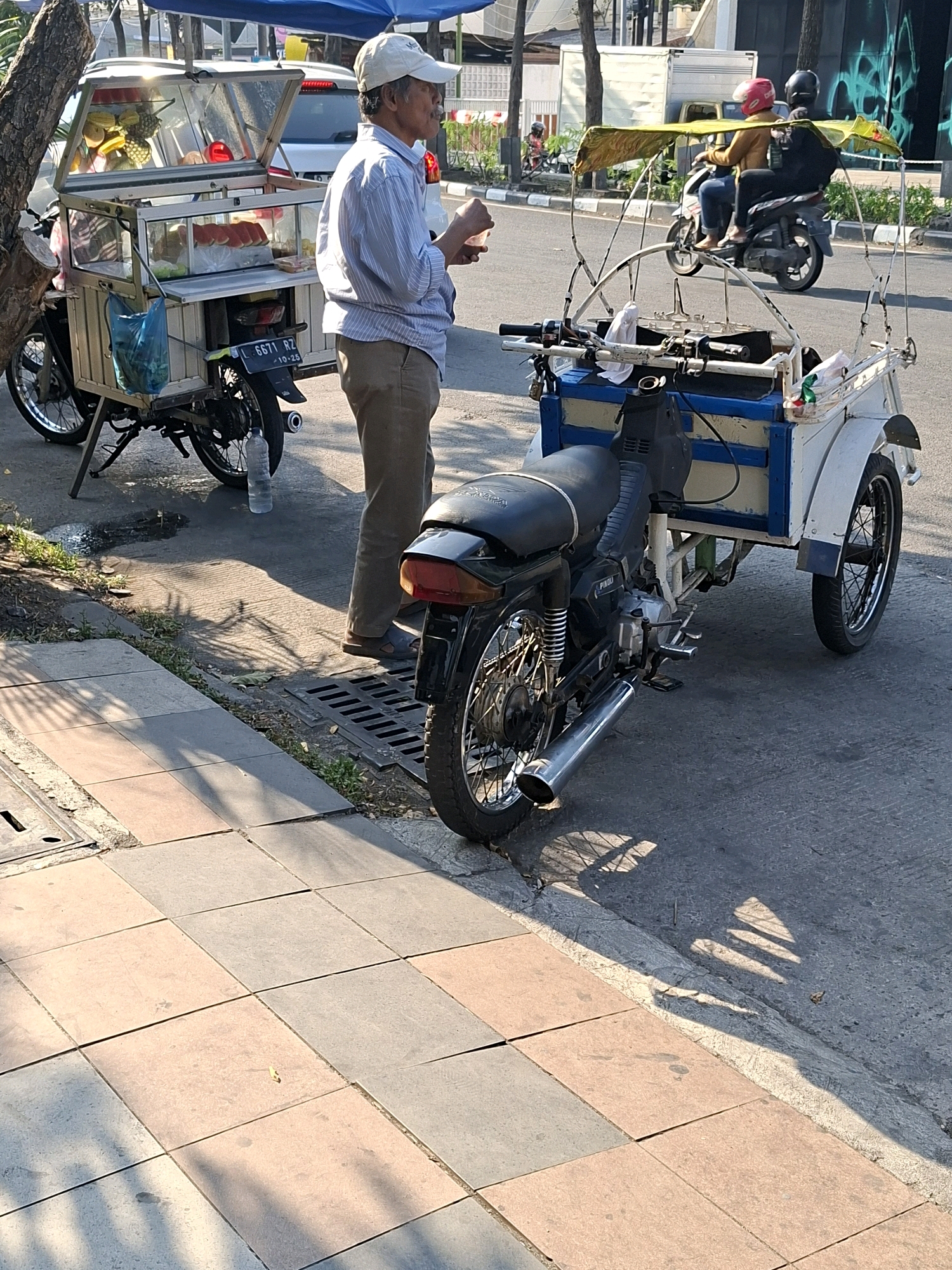 The image captures a street scene in Surabaya, Indonesia, focusing on a food vendor operating from a modified motorcycle cart. The vendor, an elderly man wearing a light blue striped shirt, beige pants, and a white cap, stands beside his cart, which is laden with various food items. The cart has a glass display case showcasing fruits, bread, and what appears to be prepared meals. A blue plastic bag hangs from the side of the cart. The motorcycle itself is a dark color, with the cart attached behind it.
In the background, two individuals are seen riding a motorcycle. The person in front wears a yellow jacket and a red helmet, while the person behind wears a black jacket and a black helmet. They are proceeding down the street, which is paved and shows signs of traffic. The environment suggests an urban setting with trees lining the sidewalk and buildings in the distance. The sun casts strong shadows on the ground, indicating it is likely daytime. The sidewalk is tiled with square pavers in alternating shades of pink and grey. A metal grate is visible on the sidewalk near the vendor's feet. A plastic water bottle is positioned on the ground near the motorcycle's rear wheel.