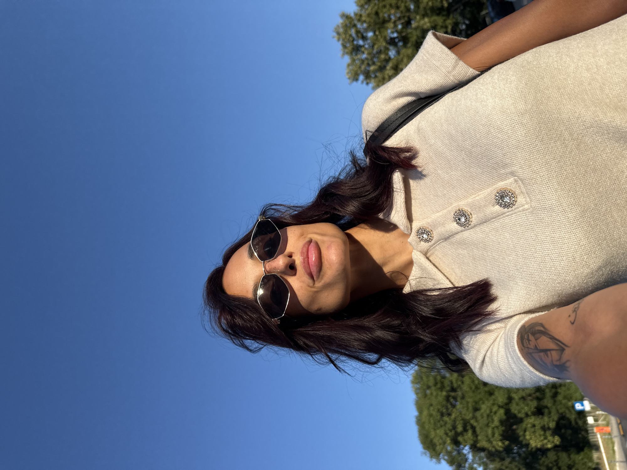 The image is a close-up, low-angle, outdoor, selfie-style shot of a woman with dark hair and sunglasses against a clear blue sky. She is wearing a light-colored collared shirt with decorative buttons. Part of her arm with a tattoo is visible on the left. Trees are in the background, and a blurry road and some signage are visible in the lower left corner. The lighting suggests it is daytime, likely late afternoon due to the warm, golden light hitting her face. There is no visible indication of the location being the city of Pleven, Bulgaria.
