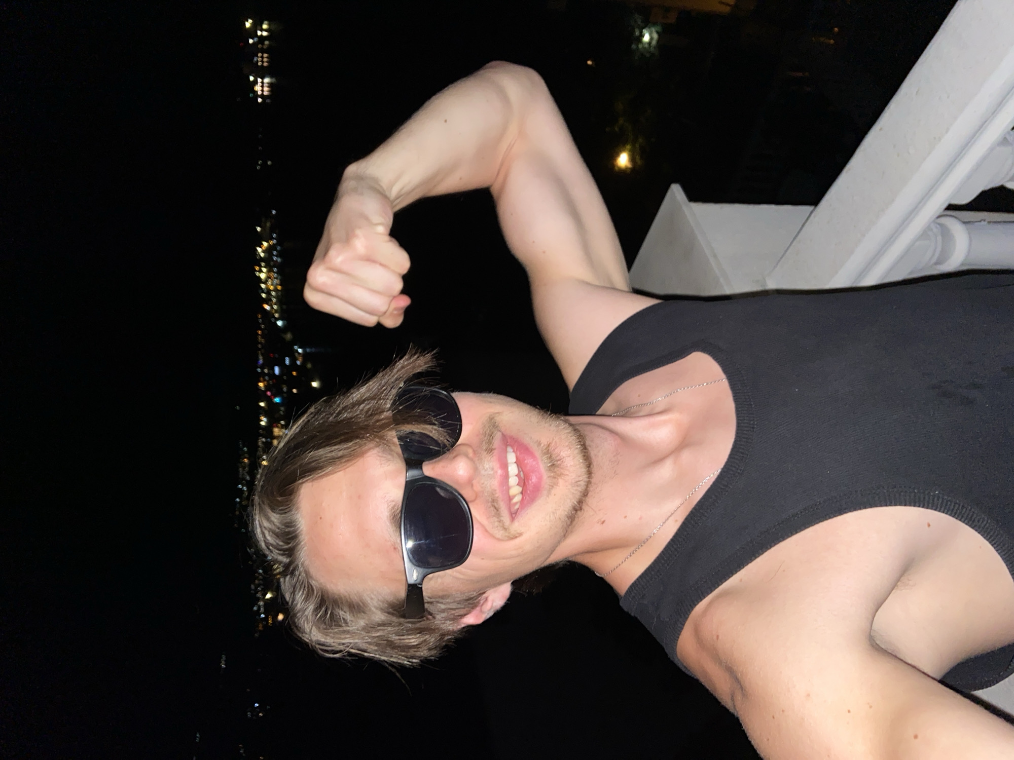 A man stands on a balcony at night in Šušanj, Montenegro. The lights of the city are visible in the background. The man is wearing sunglasses and a black tank top. He has his arm flexed, showing his bicep. A white railing is visible on the right side of the frame. The man is smiling. A silver chain is around the man's neck.