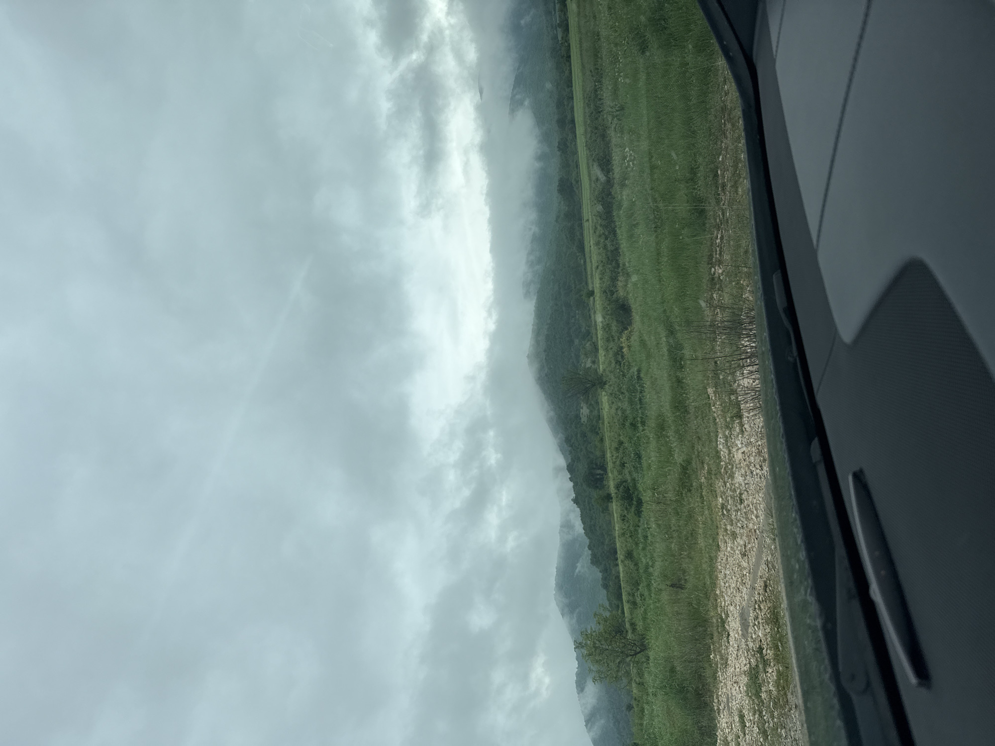 The image shows a view from inside a vehicle, likely a car. Outside the window, there is a landscape with a sky filled with gray clouds. A mountain partially obscured by mist is visible. The ground near the mountain is grassy, with some rocky areas closer to the viewer. This may be the edge of the city of Kliment in Bulgaria.