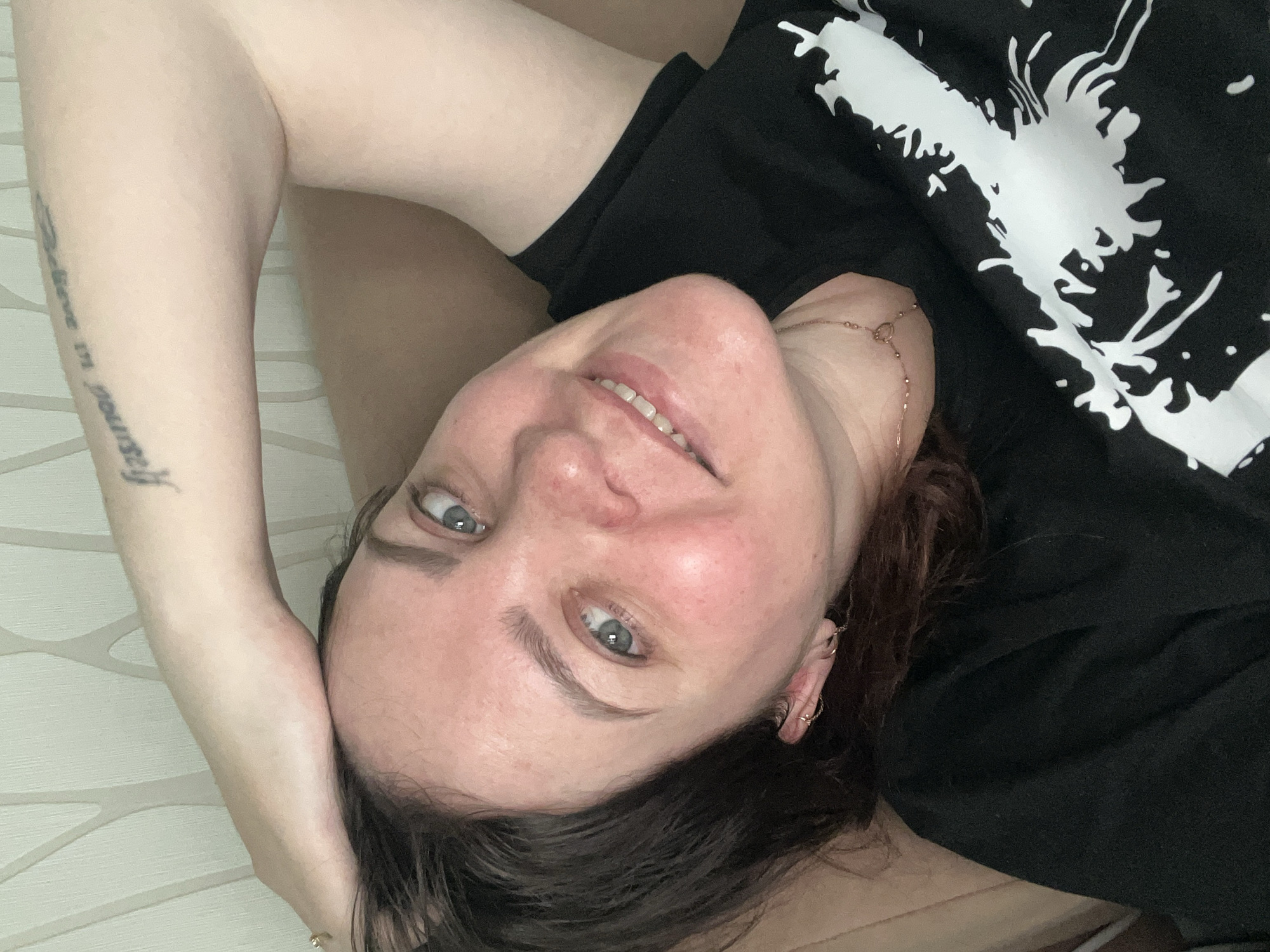 A close-up portrait features a woman lying down and looking directly at the camera with a soft, friendly smile. She has dark hair, striking blue-grey eyes, and fair skin with a slight redness visible on her cheeks and nose. She appears relaxed and content.
The woman is wearing a black t-shirt featuring a prominent white graphic design that resembles an abstract splash or splattered paint. A delicate gold chain necklace with a small pendant is visible around her neck, and she has small hoop earrings in her left ear. Her left arm is bent, with her hand positioned behind or beside her head. On her left forearm, a tattoo is clearly visible, spelling out the phrase "Believe in yourself" in a decorative script.
The background is a light-colored wall adorned with a subtle, repeating geometric pattern, suggesting an indoor setting, likely a home or private room. The lighting is soft and ambient, indicating it is daytime without direct harsh sunlight. The image captures a casual and intimate moment. This scene is set in Perm, Russia.