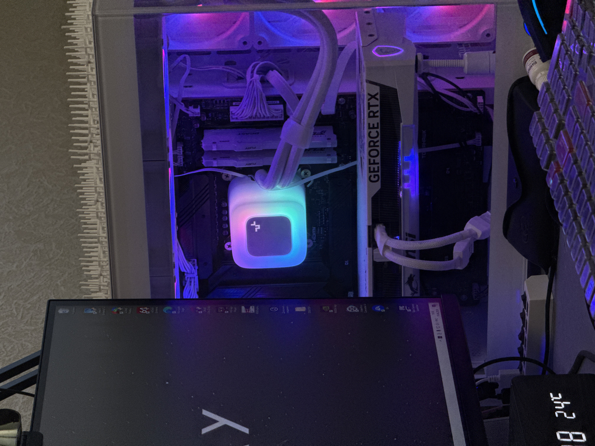 This media file depicts a close-up, overhead view of a computer setup. The primary focus is on the internal components of a desktop computer, illuminated by colorful RGB lighting, creating a vibrant and modern aesthetic. Visible inside the computer case are a graphics card labeled "GEFORCE RTX," RAM modules with "FURY" branding, and a CPU cooler with a glowing, multi-colored light. Several white cables are neatly organized, connecting the various components.
In the foreground, a portion of a monitor is visible, displaying desktop icons and a wallpaper with a stylized "Y." To the right of the computer case, a portion of a keyboard with RGB lighting can be seen, along with a small digital display showing "38 24C." The overall scene suggests a gaming or high-performance computing environment. The lighting creates a somewhat dark ambiance, with the computer's illumination being the dominant light source.
There are no people or explicit weather or time of day cues, though the lighting suggests it might be evening or night. The presence of computer hardware and a monitor indicates an indoor setting, likely a home office or gaming room. The text visible includes brand names and model numbers of computer components, as well as icons and text on the monitor's desktop. The location context provided is Shushary, Russia.