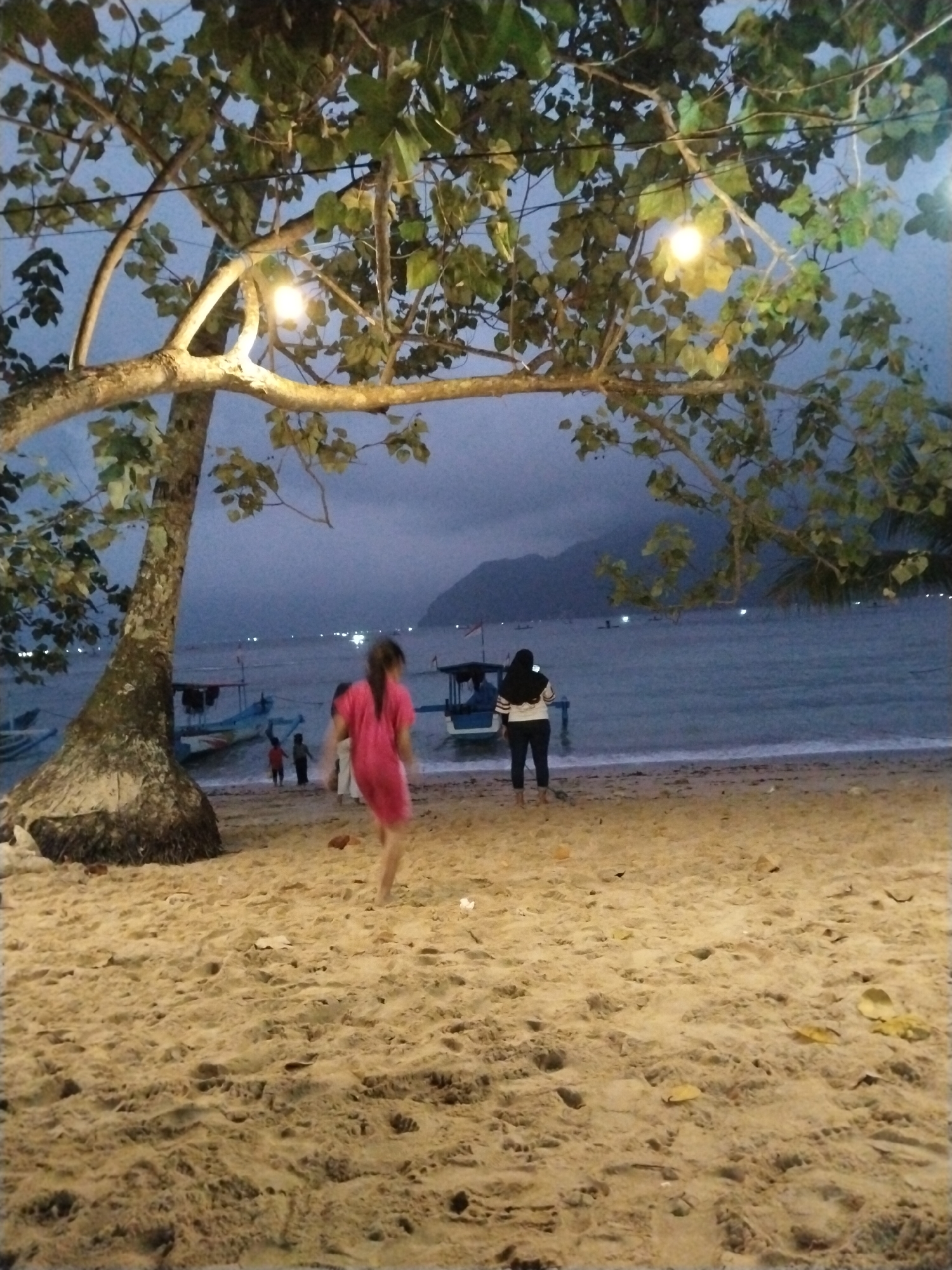 The image captures a dimly lit beach scene at dusk, likely in Prigi, Indonesia, as suggested by the overall ambiance and proximity to a body of water with boats. A large tree with its branches extending into the frame dominates the foreground, with a few warm lights visible among the leaves. The sandy beach is textured with footprints and scattered leaves. In the middle ground, several small boats are beached or partially submerged in the water. A few people are present. In the left-center, a person in a bright pink dress is walking away from the camera towards the water. To the right, a person wearing a black hijab and a white and black striped top stands facing the water. Further back, near the boats, a few other individuals are visible, their forms less distinct due to the low light. The background shows a body of water, with distant lights suggesting a shoreline or other boats. Mountains are faintly visible on the horizon, shrouded in twilight.